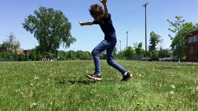 Un enfant joue dans un parc, sautant ou dansant sur l'herbe sous un ciel bleu clair.