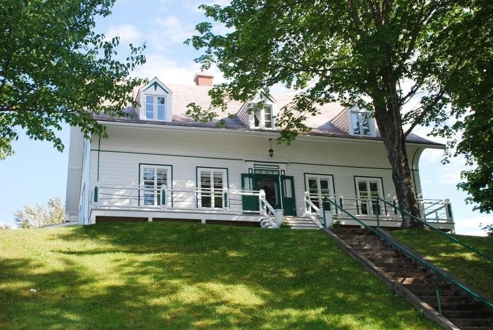 Maison blanche avec balustrades sur la terrasse, entourée d'arbres et située en haut d'une pente herbeuse avec un escalier en dessous.