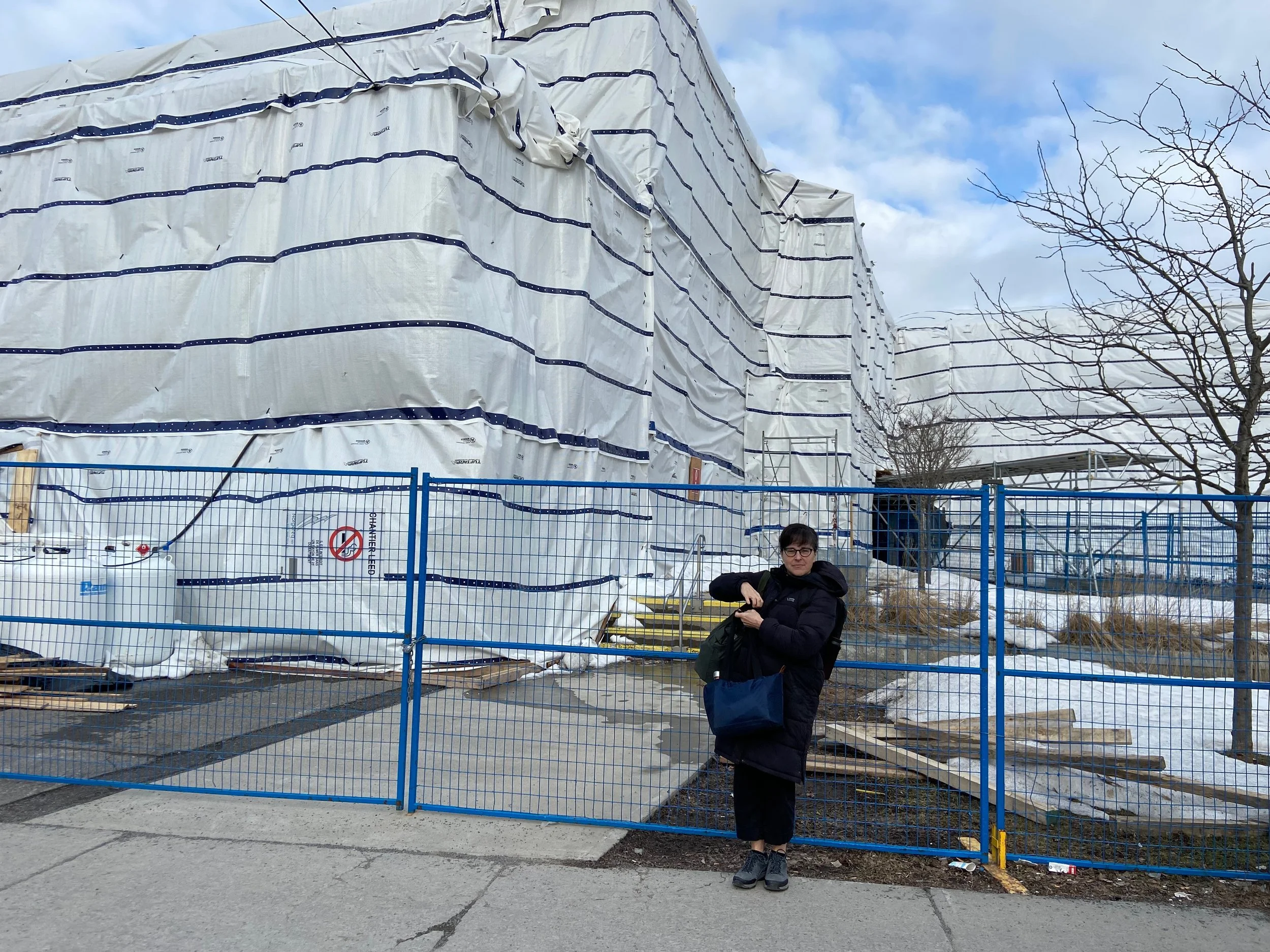 Une personne debout devant une clôture en métal bleu, portant un manteau noir, des lunettes, tenant un sac noir et un sac bleu, en face d'un bâtiment en construction ou en rénovation qui est recouvert de bâches blanches avec des lignes bleues, dans un environnement en extérieur avec des arbres sans feuilles et un ciel nuageux, en hiver.