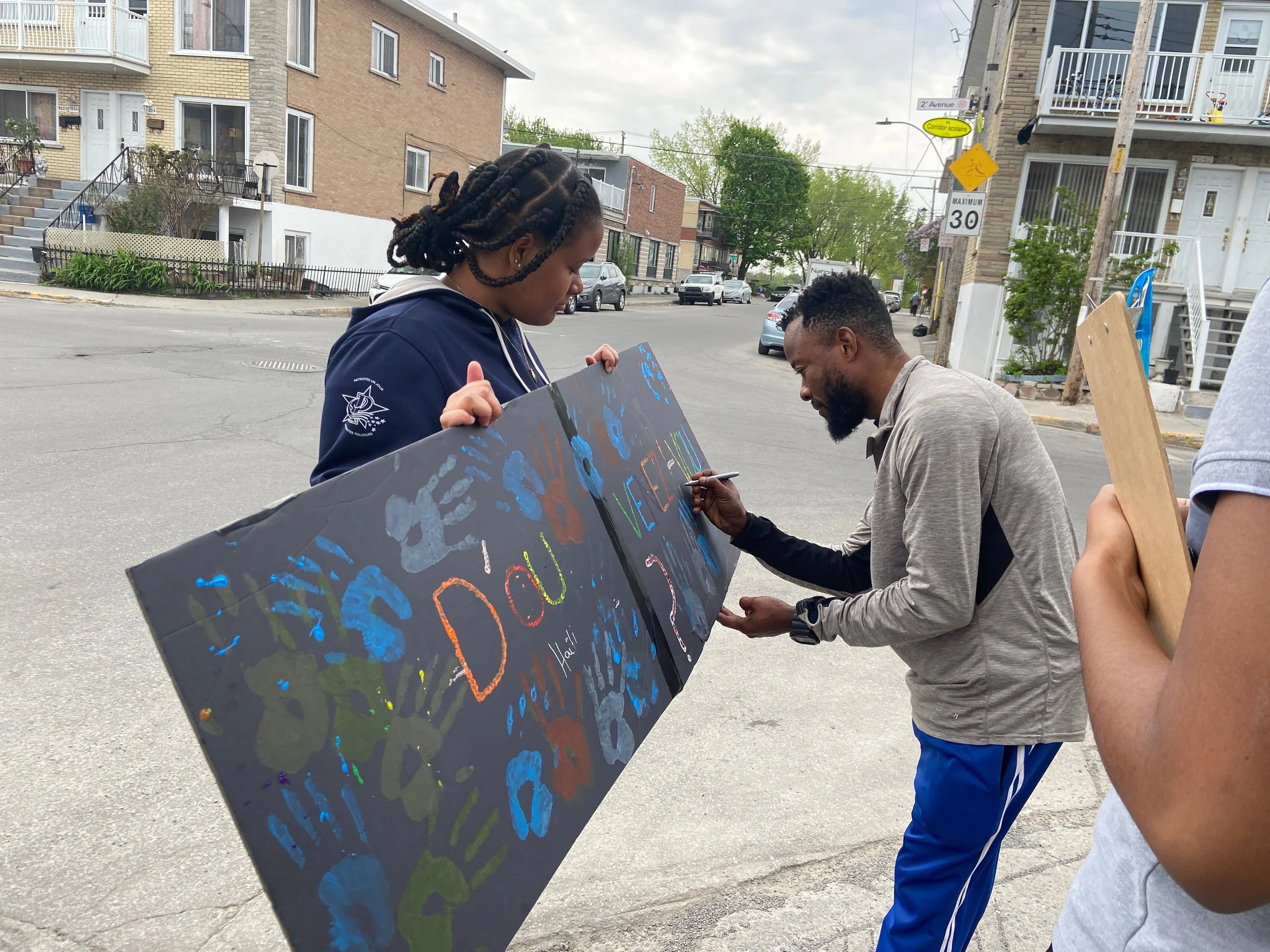 Un homme et une femme tiennent un panneau d'art dans la rue, avec des mains peintes en différentes couleurs, et ils participent à une activité artistique ou de sensibilisation.