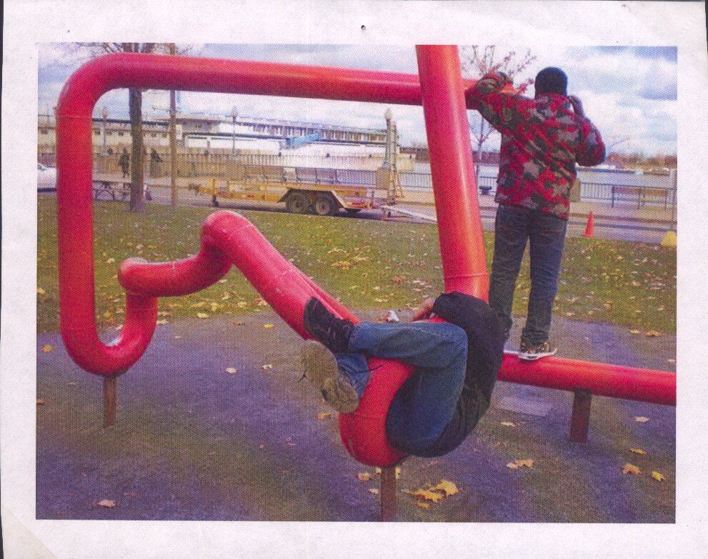 Deux enfants jouent sur un parcours de jeux urbain en métal rouge dans un parc, avec un fond de bâtiment et de ciel nuageux.