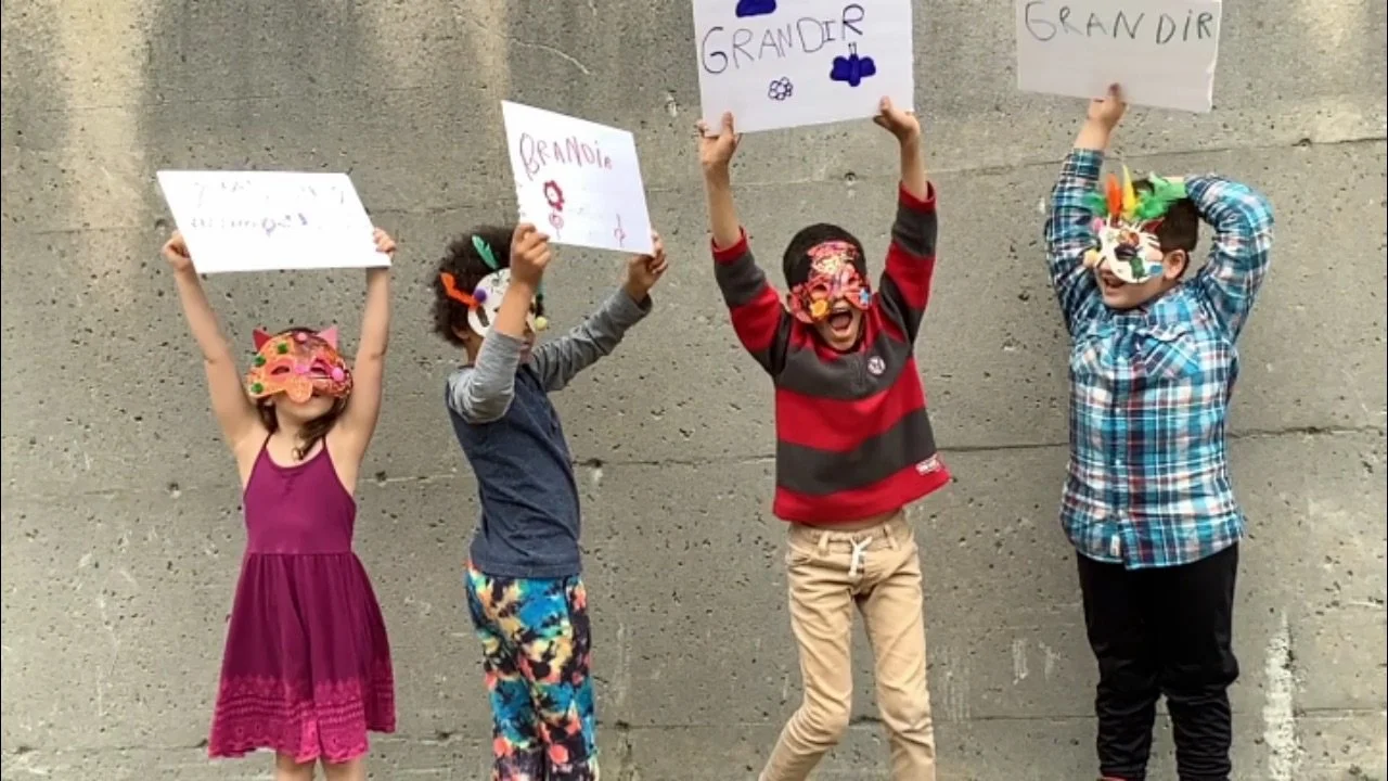 Quatre enfants portant des masques colorés et tenant des panneaux avec des mots comme "Grandir" et "Grande", se tenant contre un mur en béton, célébrant une fête ou un événement joyeux.