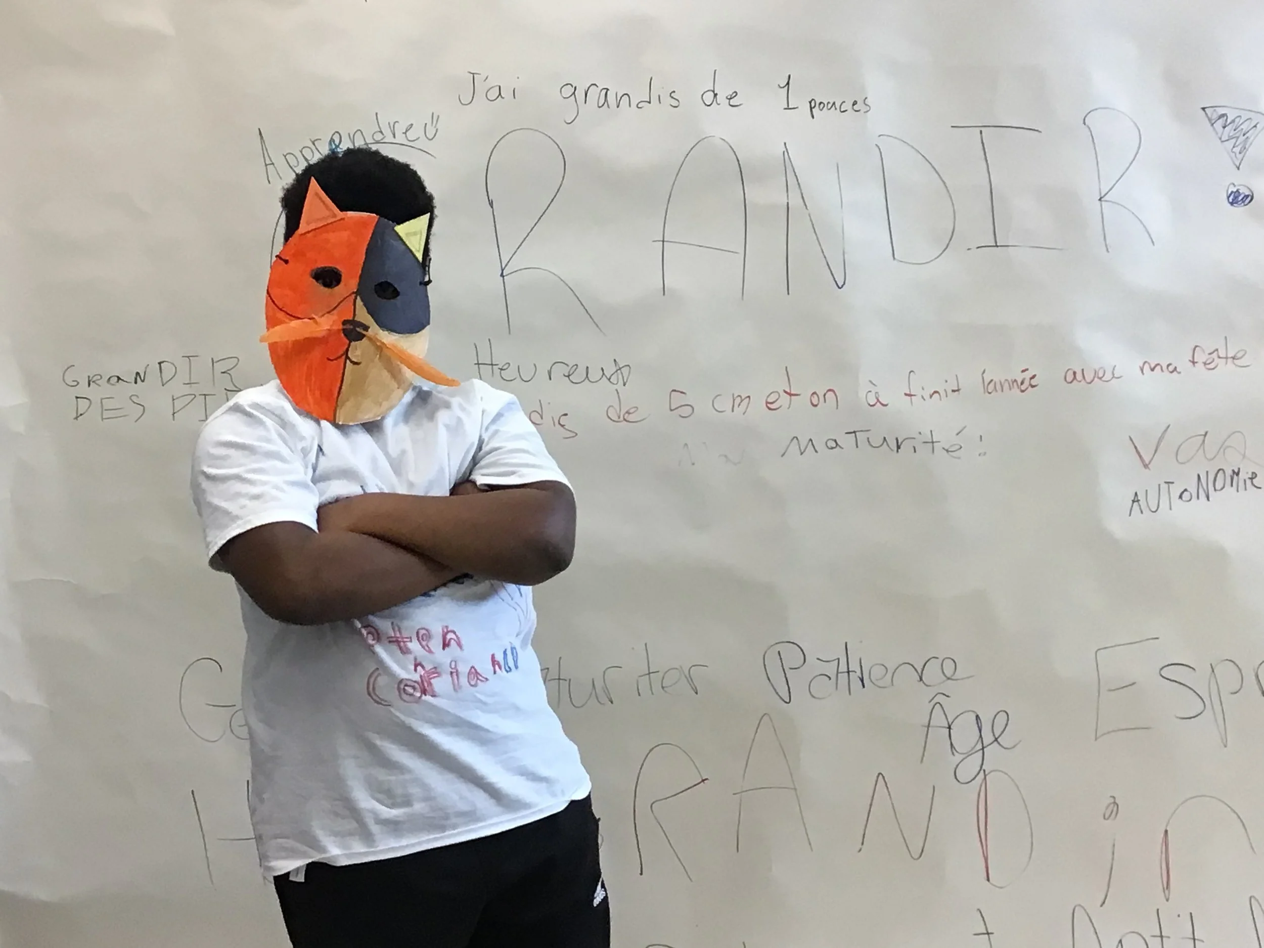 Une personne portant un masque en papier représentant un chat orange et noir, avec des oreilles en papier, se tenant devant un tableau blanc avec des écrits en français.