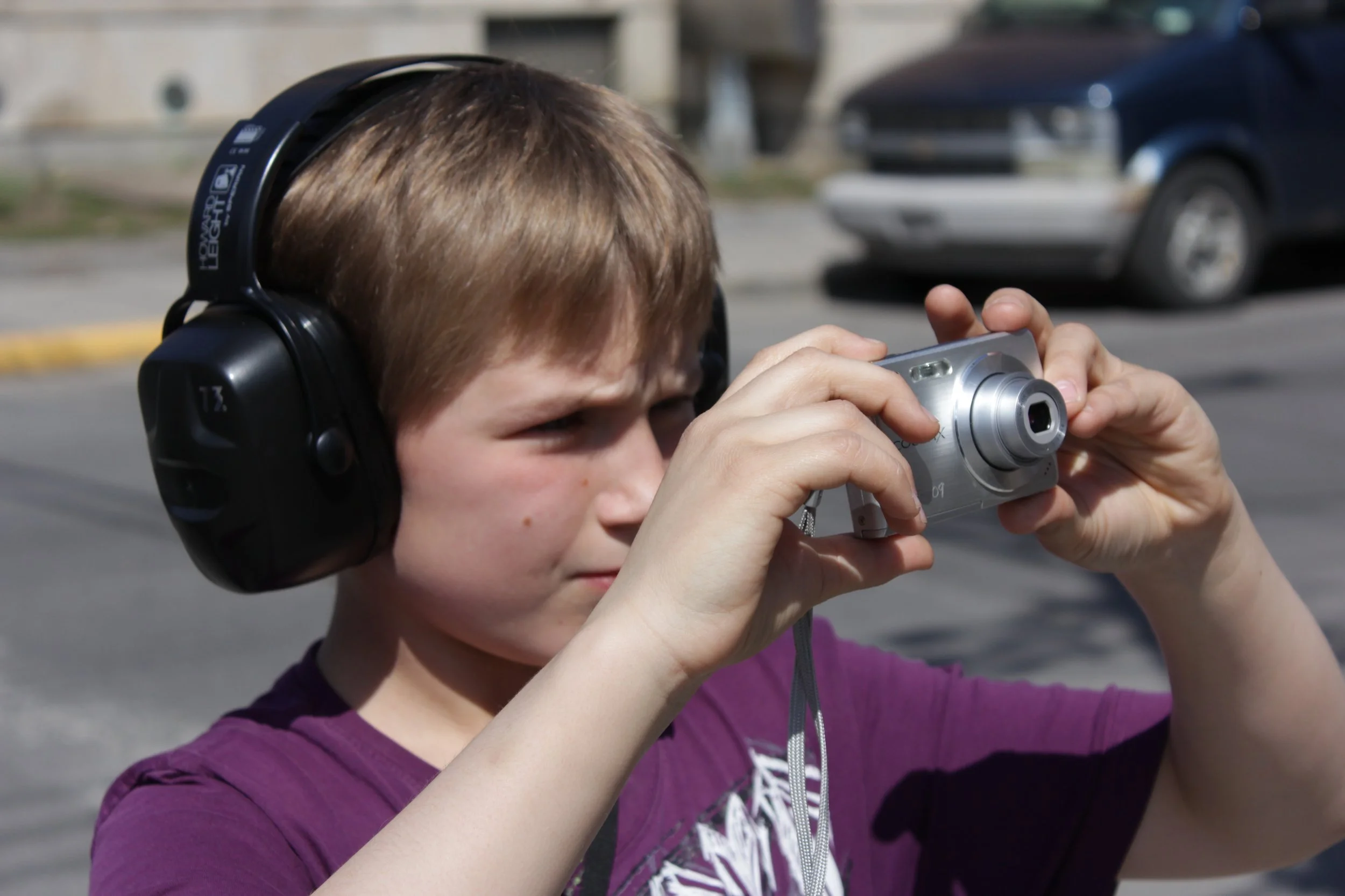 Un garçon jeune portant un casque antibruit, tenant un appareil photo argenté, en plein air avec des voitures en arrière-plan.
