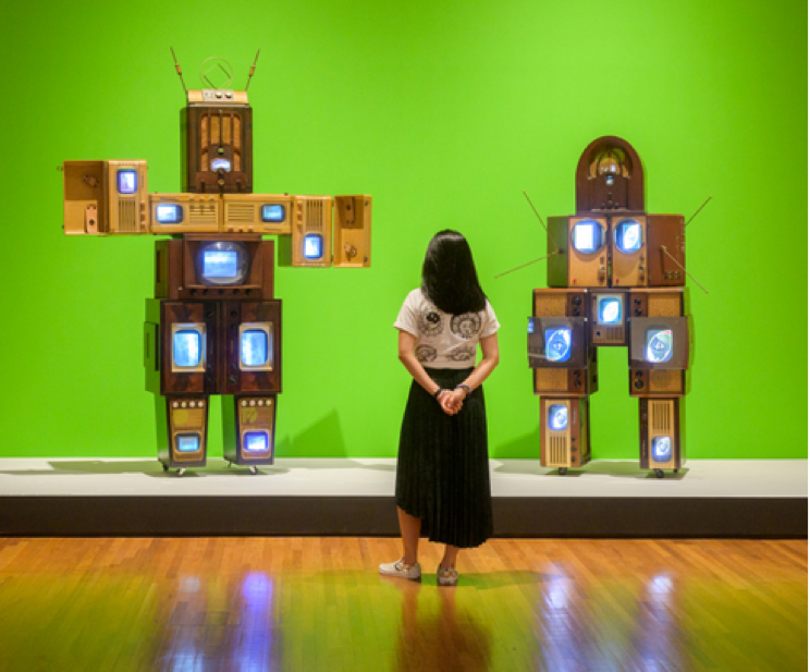 Une femme regarde des sculptures composées d'anciens téléviseurs et appareils électroniques disposés en formes humanoïdes sur un fond vert dans une galerie d'art.