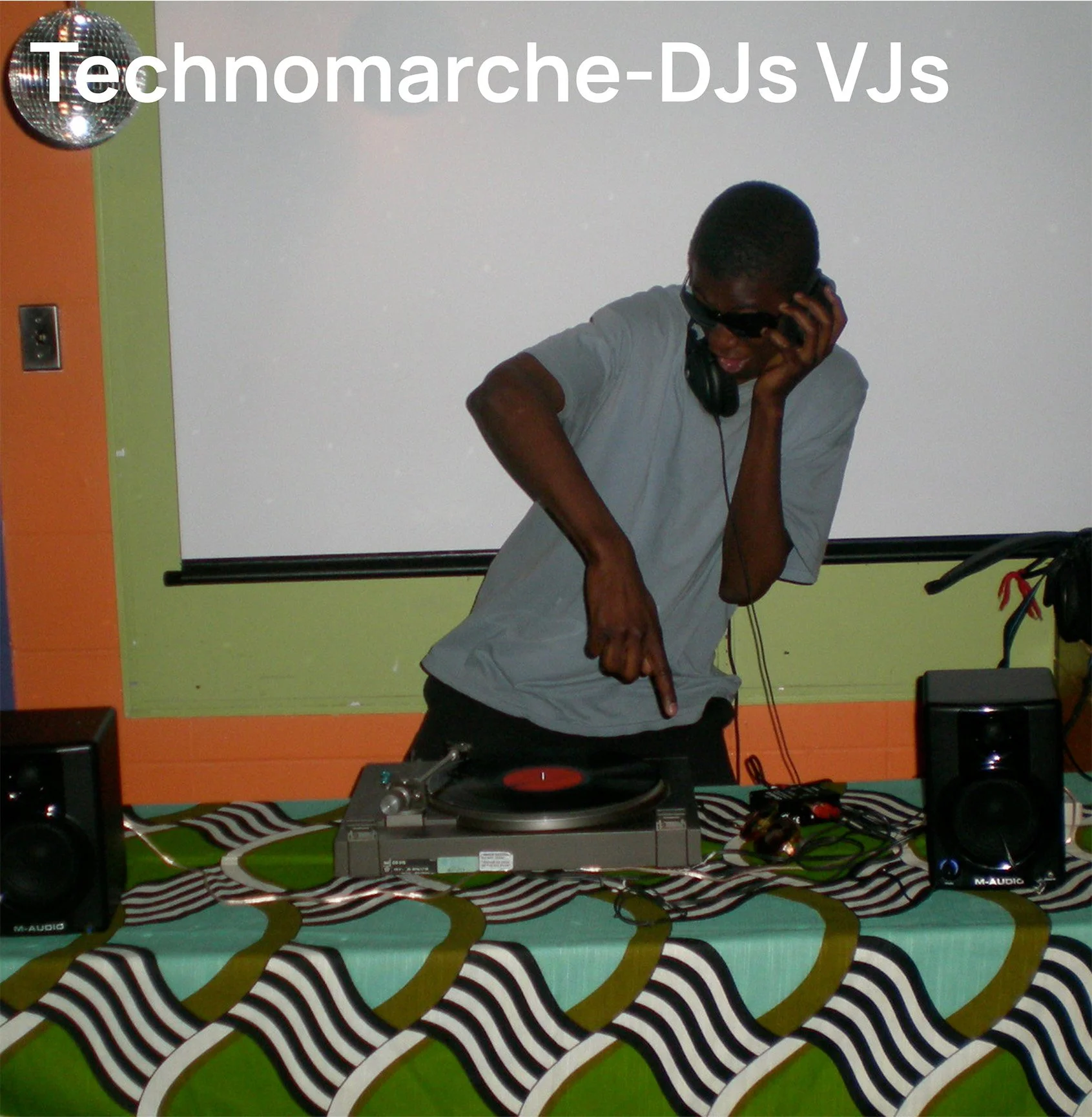 Un DJ portant des lunettes de soleil et un casque, pointant une table de mixage sur un fond de mur blanc et orange, avec un tableau blanc derrière lui et des enceintes de chaque côté.