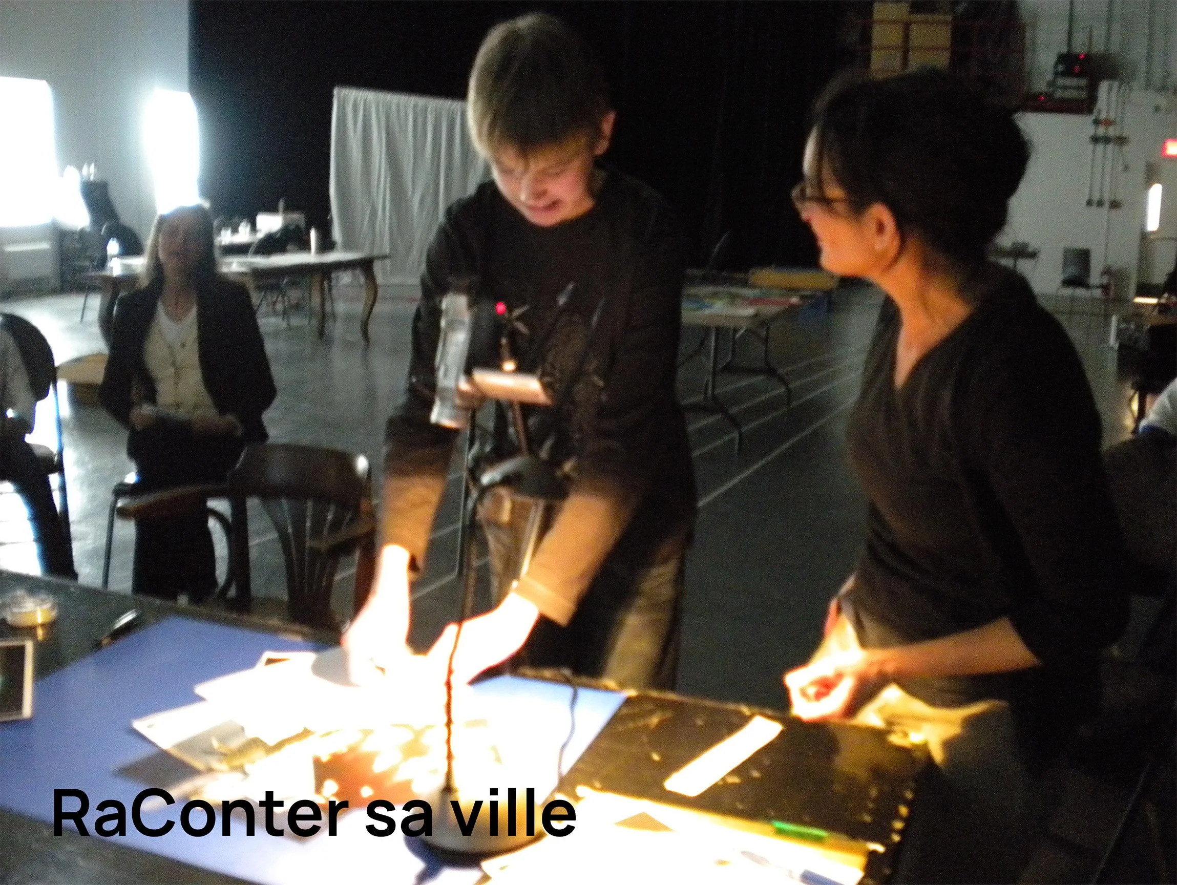 Un garçon et une femme travaillent ensemble sur un projet d'exposition ou de science, dans une pièce sombre, éclairée par une lampe de bureau. Une autre femme est assise à une distance, observant. Il semble s'agir d'une présentation ou d'une démonstr