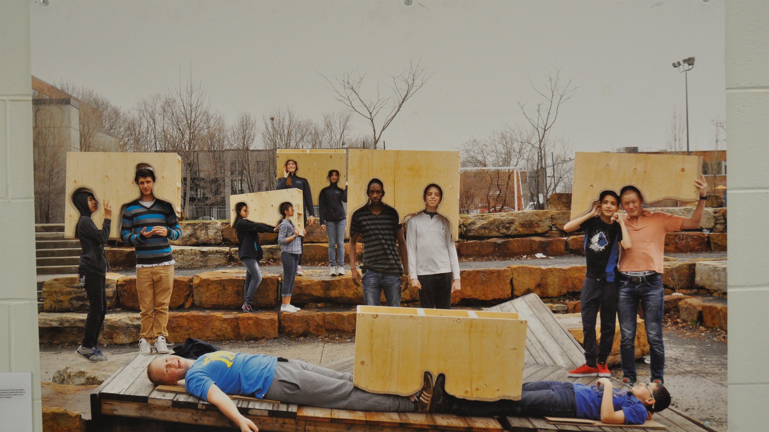 Un groupe de jeunes de différents âges pose en extérieur dans un parc, certains près d'une structure en bois avec des rochers en arrière-plan, un garçon allongé sur une plateforme en bois au premier plan, plusieurs autres debout ou assis derrière.