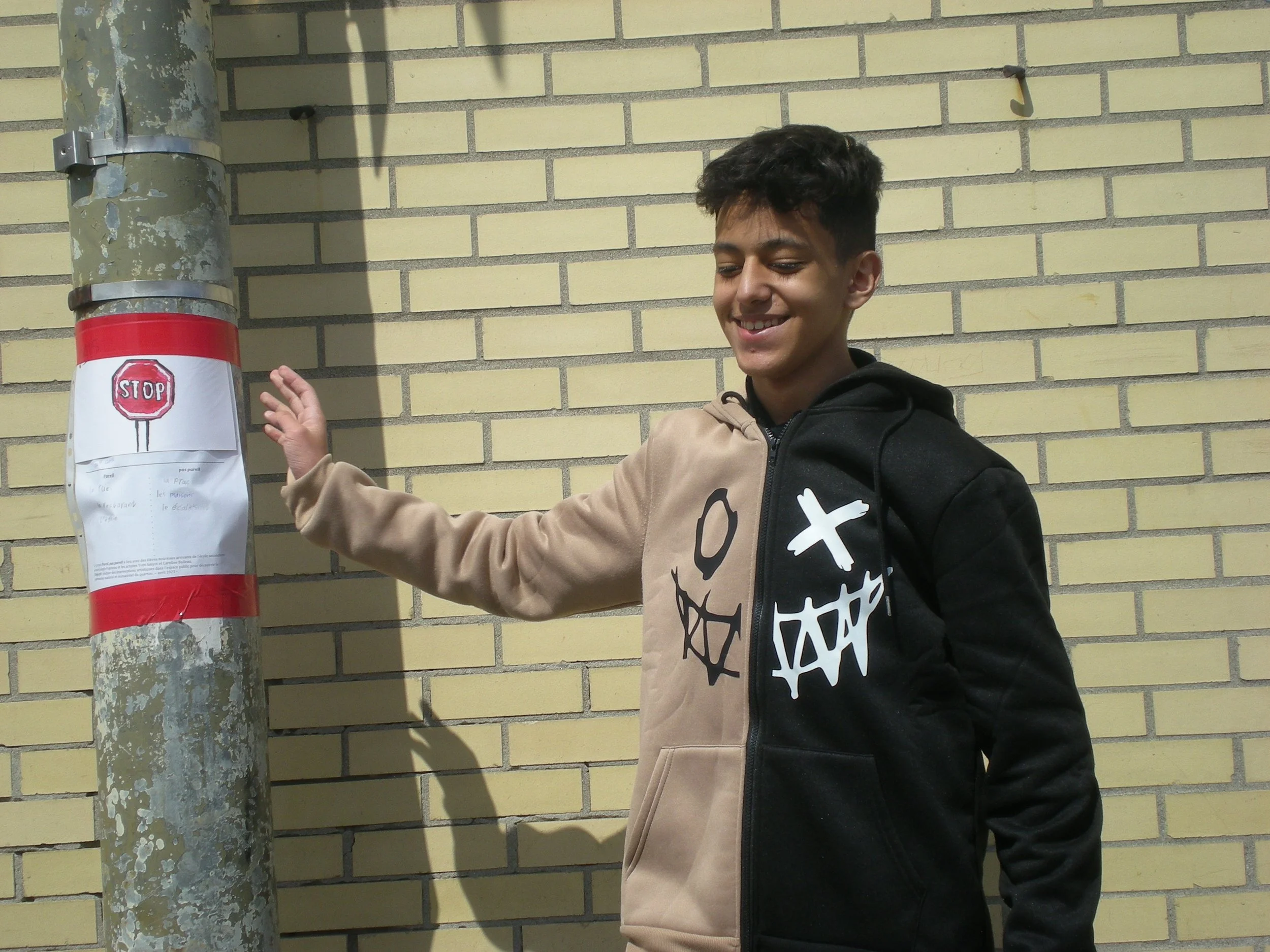 Un garçon souriant touche un panneau d'arrêt en papier collé à un poteau en métal, en présence d'un mur jaune en briques.