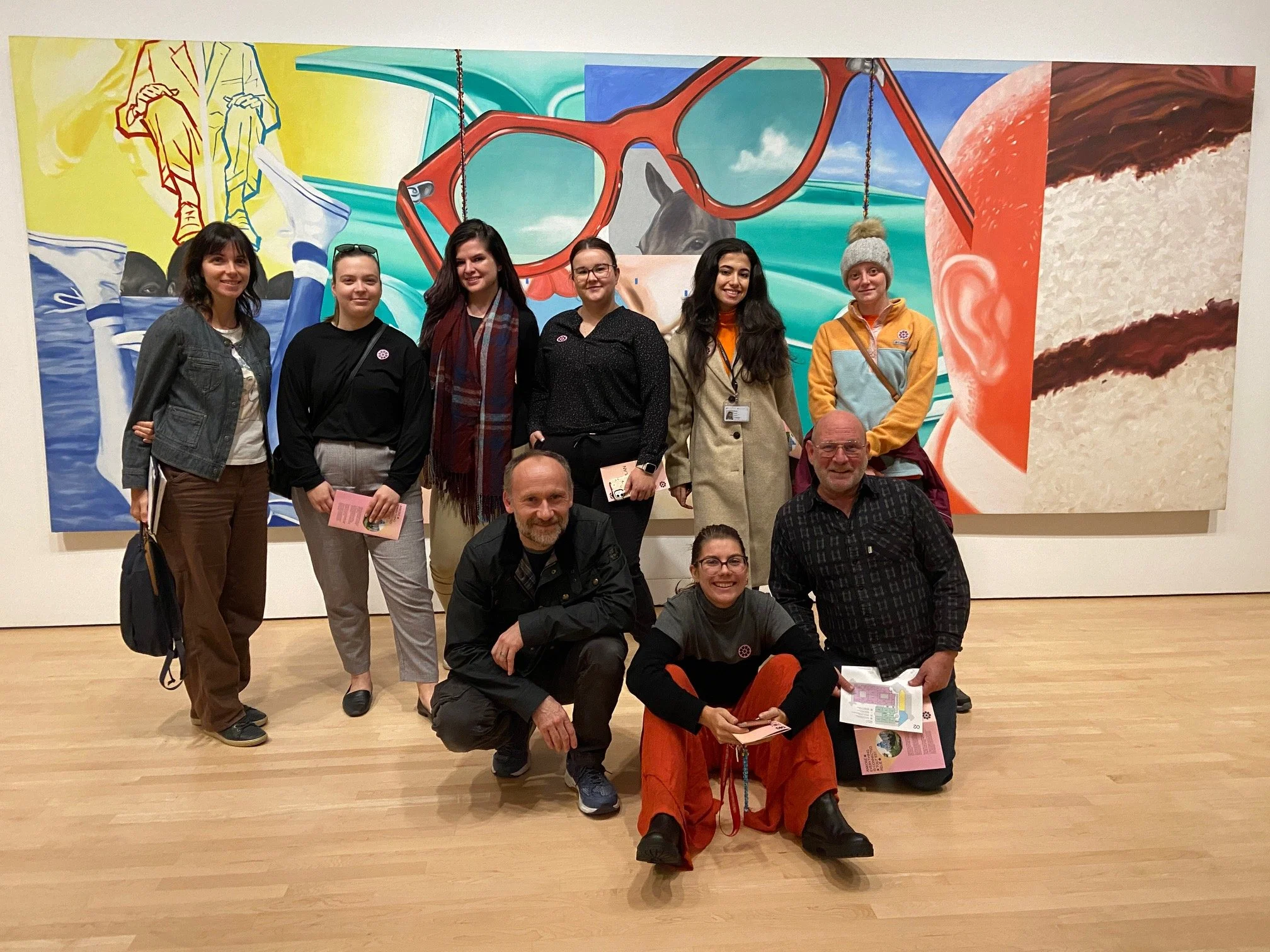 Groupe de neuf personnes posant devant une œuvre d'art colorée dans une galerie. La peinture présente des lunettes, des paysages abstraits et des éléments de style moderne.