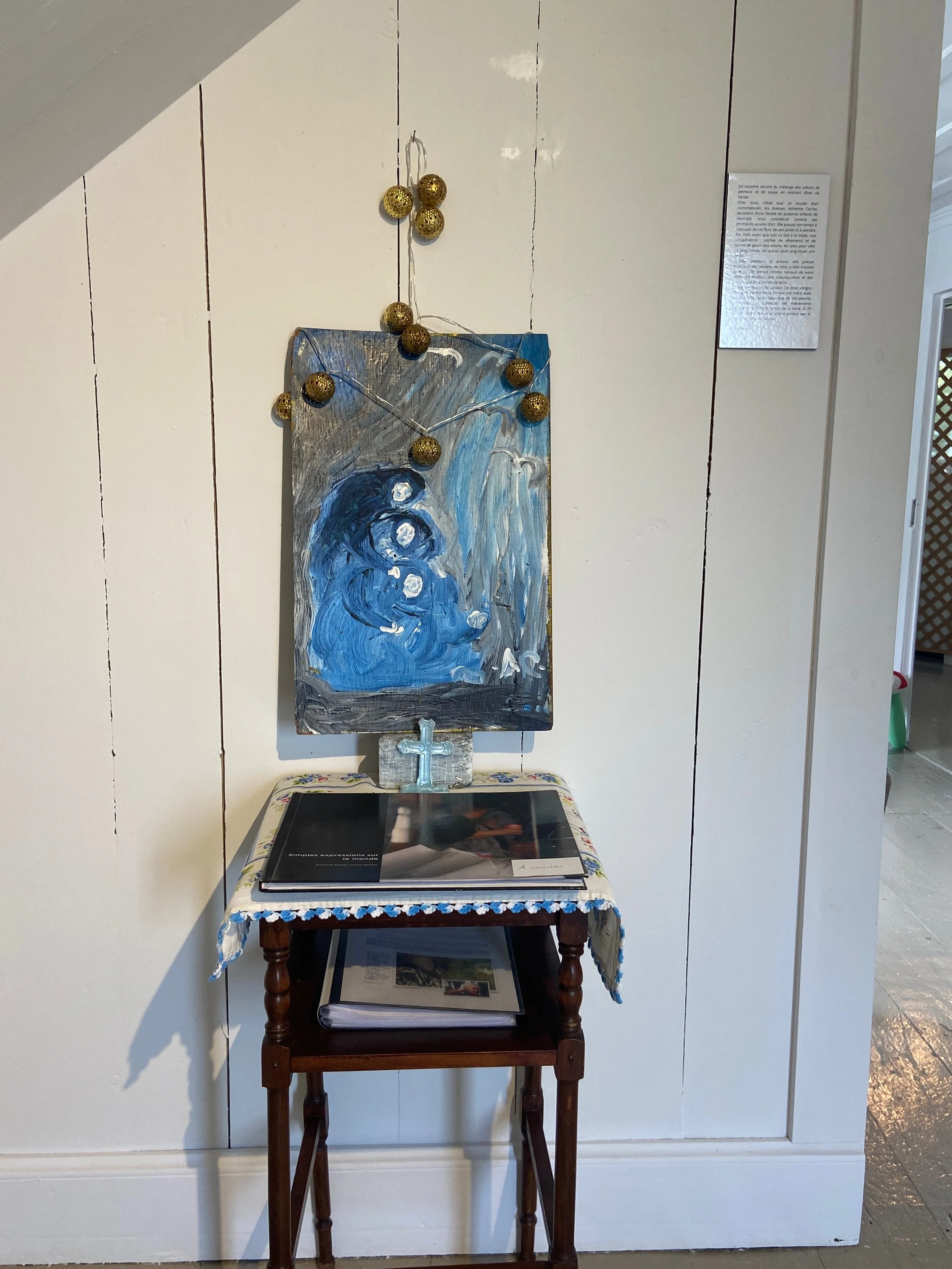 Un tableau peint à la main, une guirlande de boules dorées, une table en bois avec un livre, une croix en bois, et un autre livre en dessous, dans une pièce intérieure.