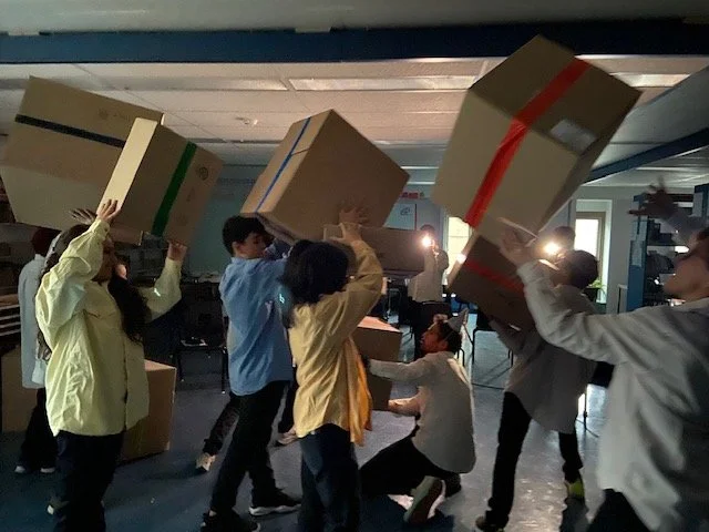 Groupe de personnes, jeunes, portant des cartons d'emballage, dans une pièce sombre, avec lumière artificielle, semblant organiser ou déplacer des objets.