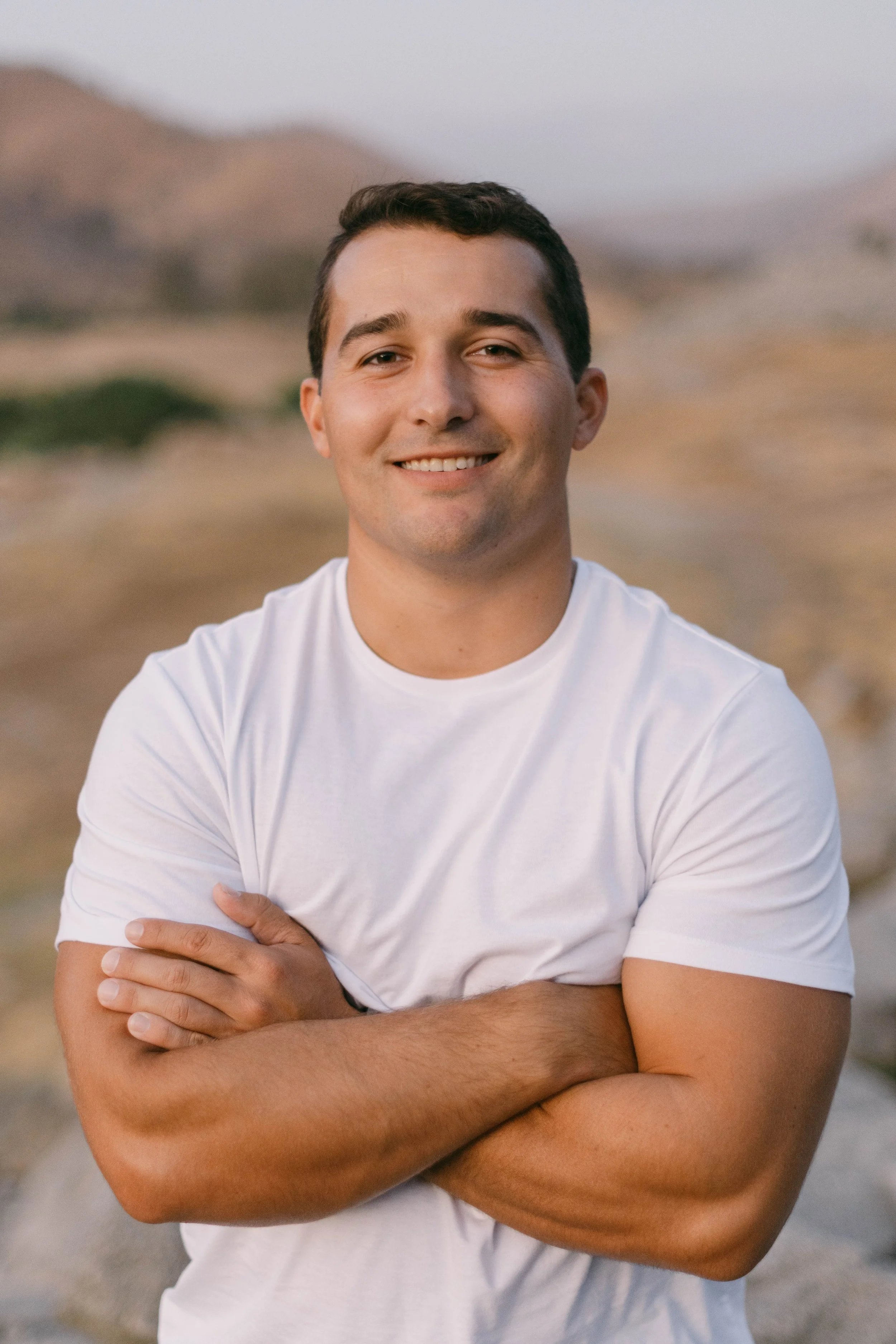 Smiling person in white t-shirt with arms crossed against a blurred natural background.