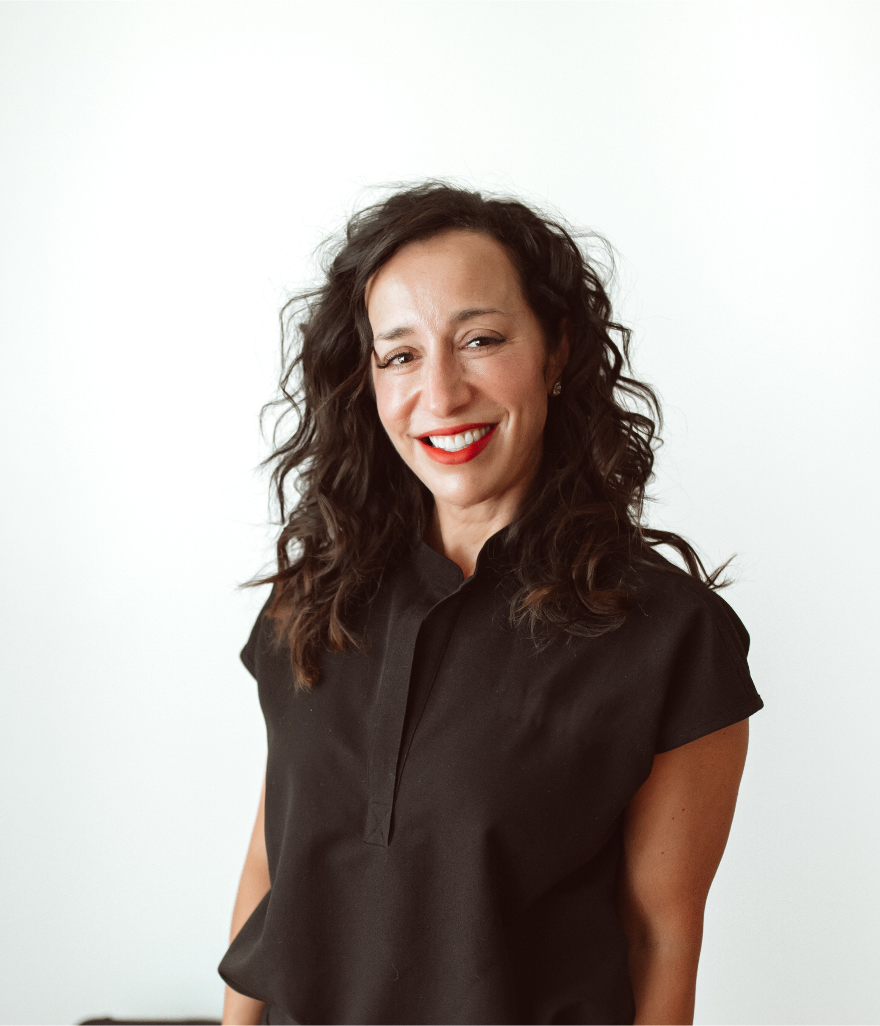 A woman with curly dark hair and red lipstick smiling at the camera, wearing a black shirt, standing against a plain white background.