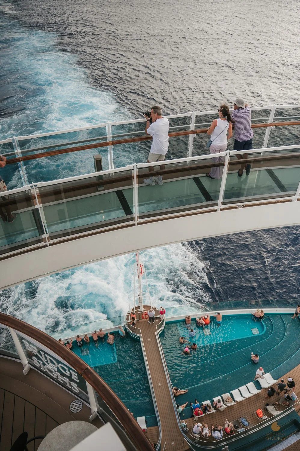 People on a cruise ship's deck, with some observing the ocean and others swimming in the pool below.