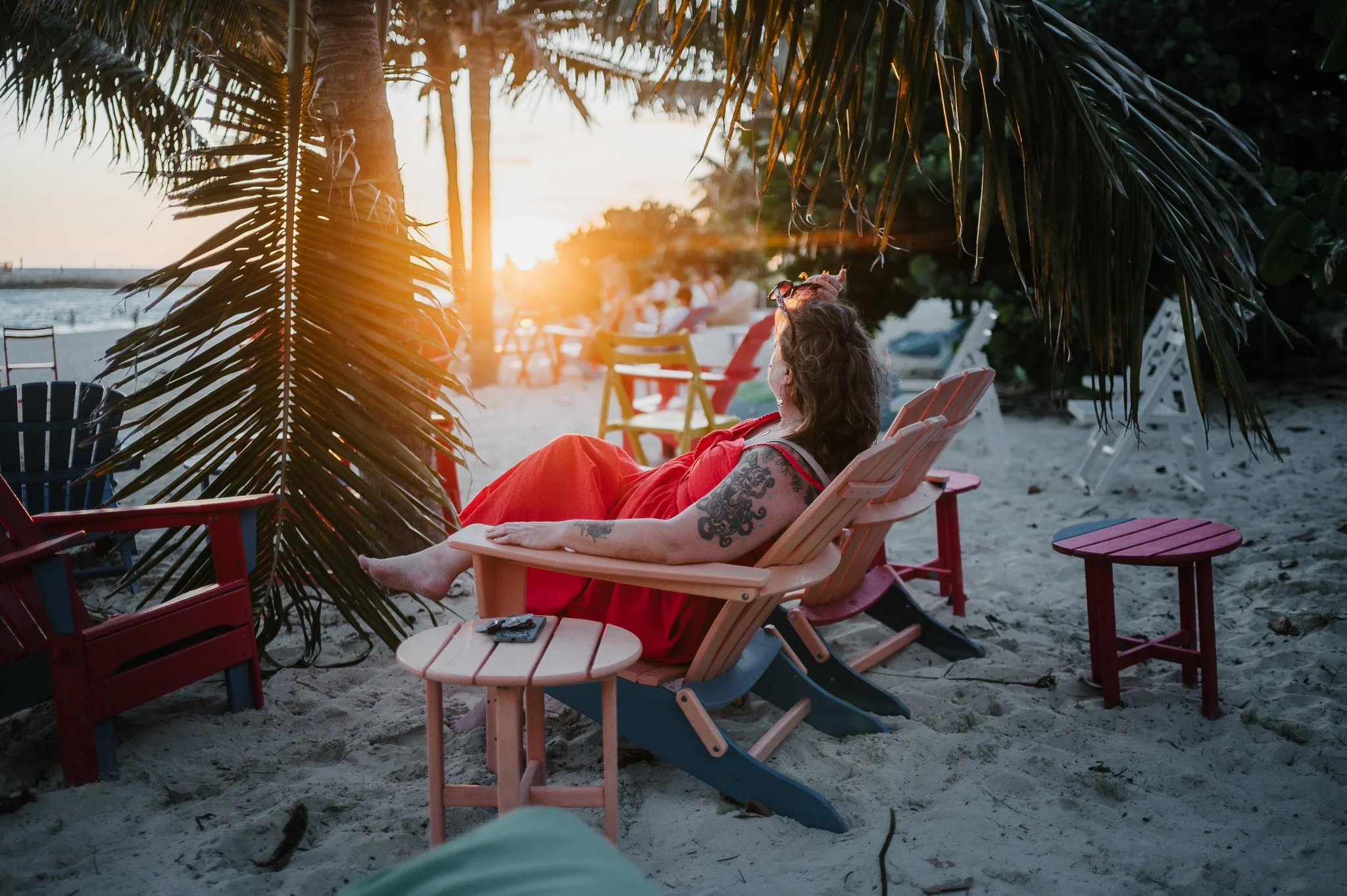 A woman with tattoos relaxing on a pink wooden beach chair, wearing a red dress, during sunset on a sandy beach surrounded by colorful chairs and palm trees.
