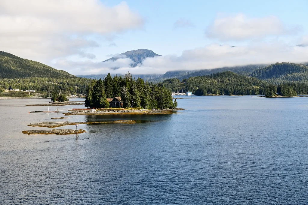 A scenic view of a body of water with a small forested island, surrounded by mountains partly covered in clouds, under a partly cloudy sky.