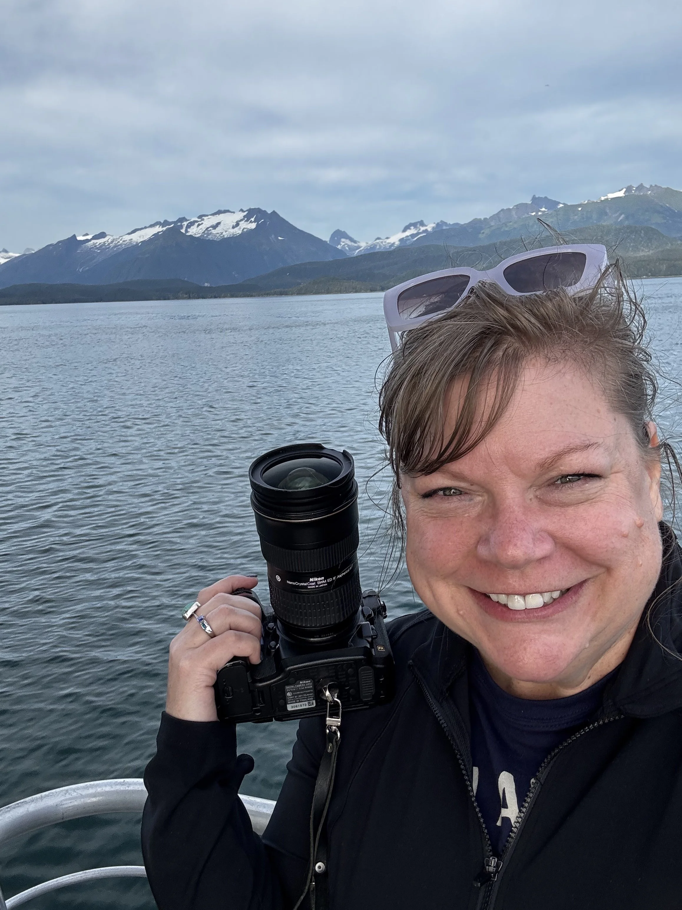 A smiling woman holding a camera with mountains and a lake in the background, wearing sunglasses on her head.