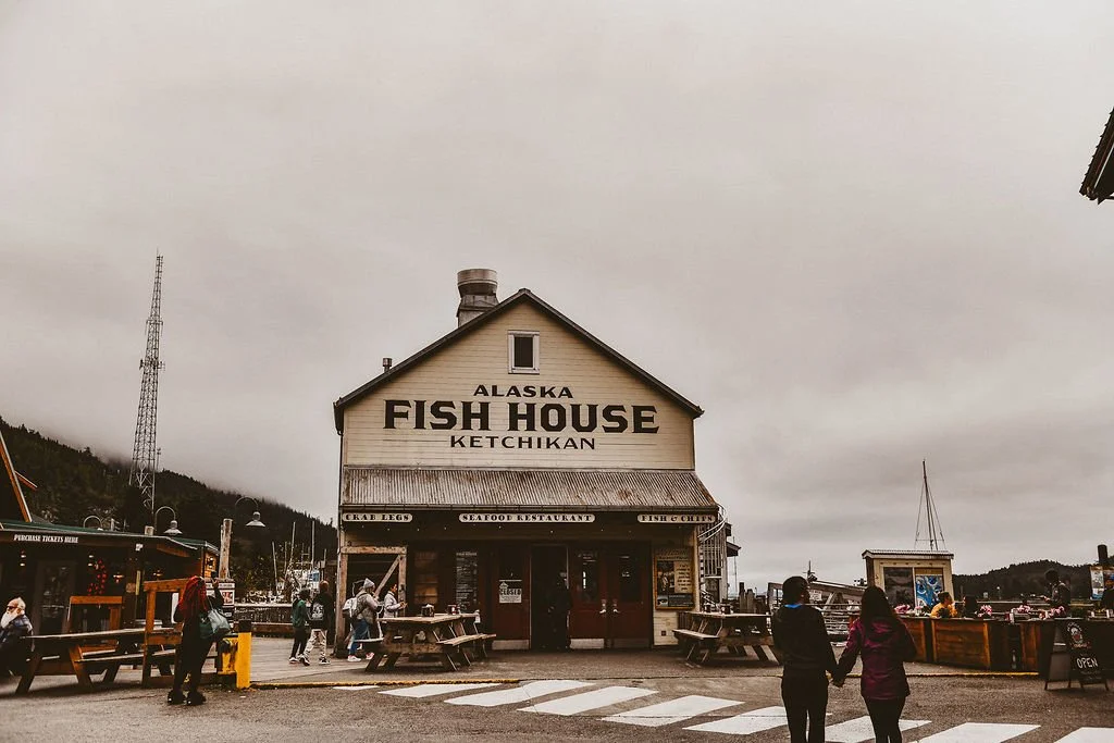 A two-story building with a sign reading 'Alaska Fish House Ketchikan' on the front. The building is light-colored with a dark roof, and it has a small window on the second story. There are people walking around and holding hands near the front, with outdoor seating and various signs indicating seafood restaurant offerings.
