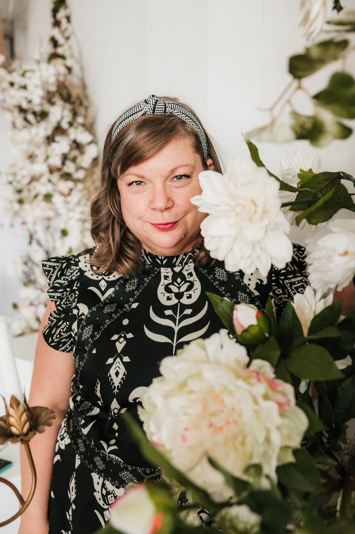 A woman with shoulder-length brown hair, wearing a black and white geometric patterned dress and a matching headband, is posing among white flowers and greenery, smiling at the camera.