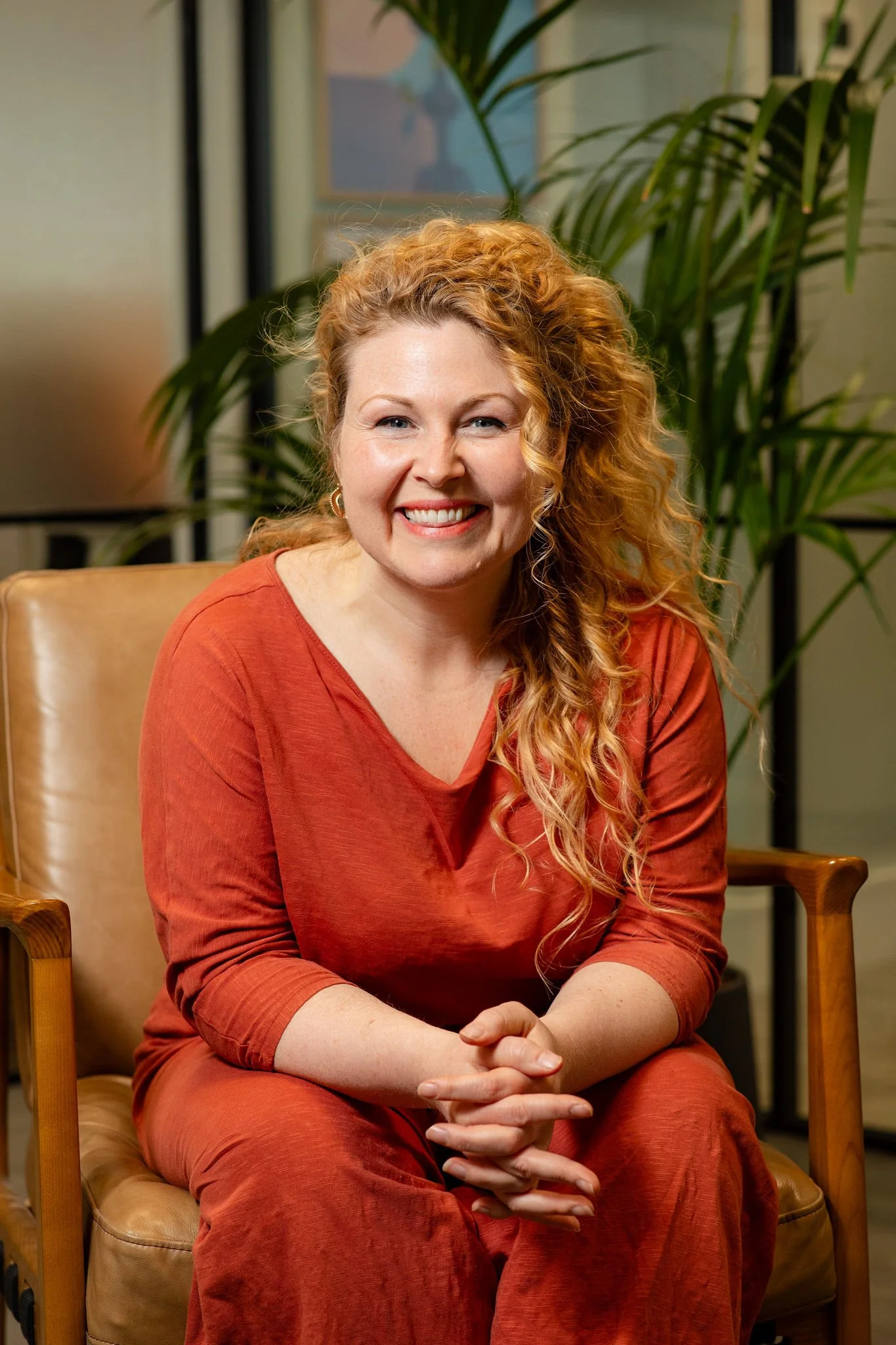 A woman with curly blonde hair, wearing a burnt orange dress, sitting on a tan chair, smiling with her hands clasped in her lap. Behind her is a green plant and a frosted glass door.