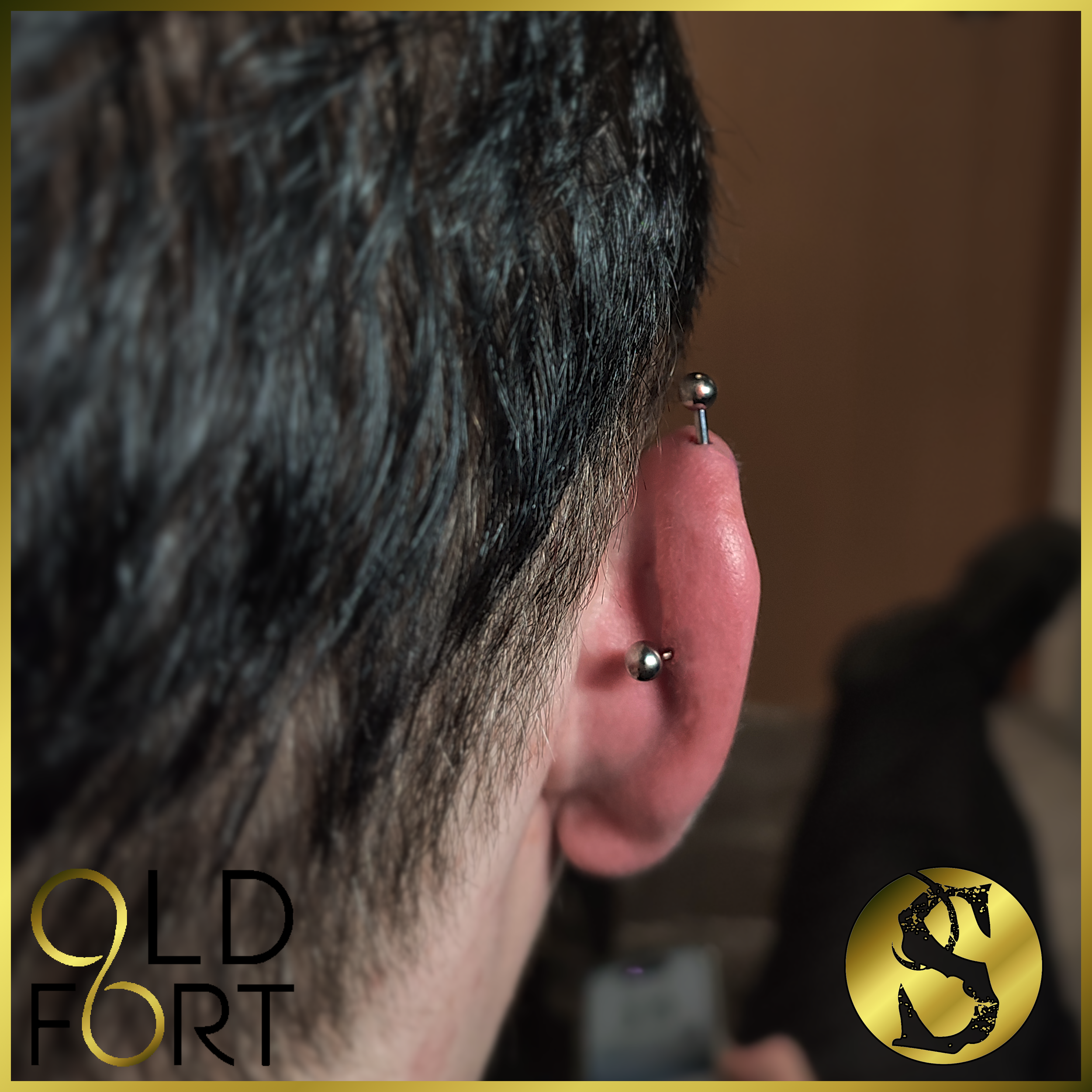 Close-up of a person's ear with two metal piercings, one on the earlobe and one on the upper cartilage, with dark hair partially covering the ear.