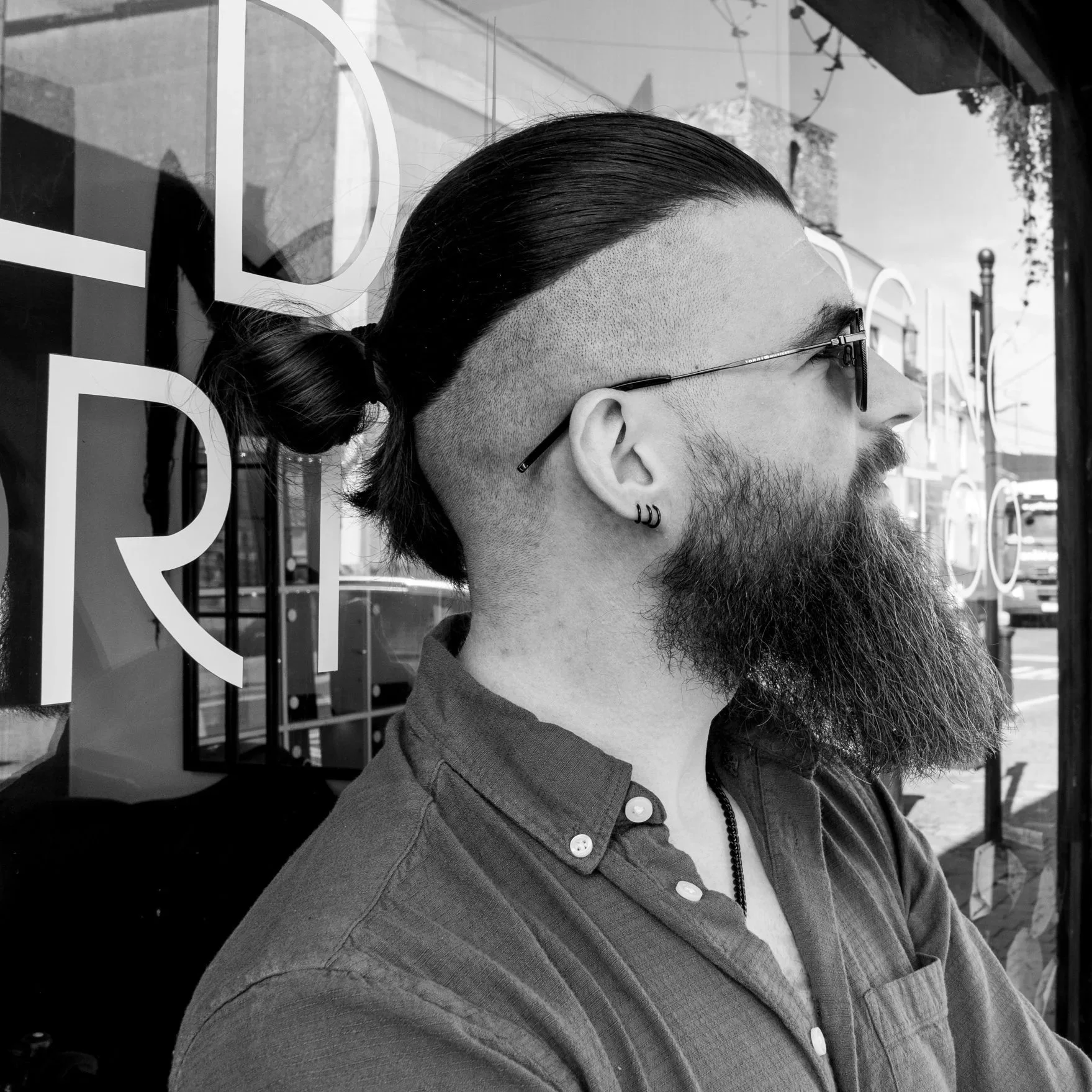 Profile of a man with a slicked-back ponytail, shaved sides, beard, wearing sunglasses, earrings, and a button-up shirt, standing outside a shop with large windows and street visible in the background.