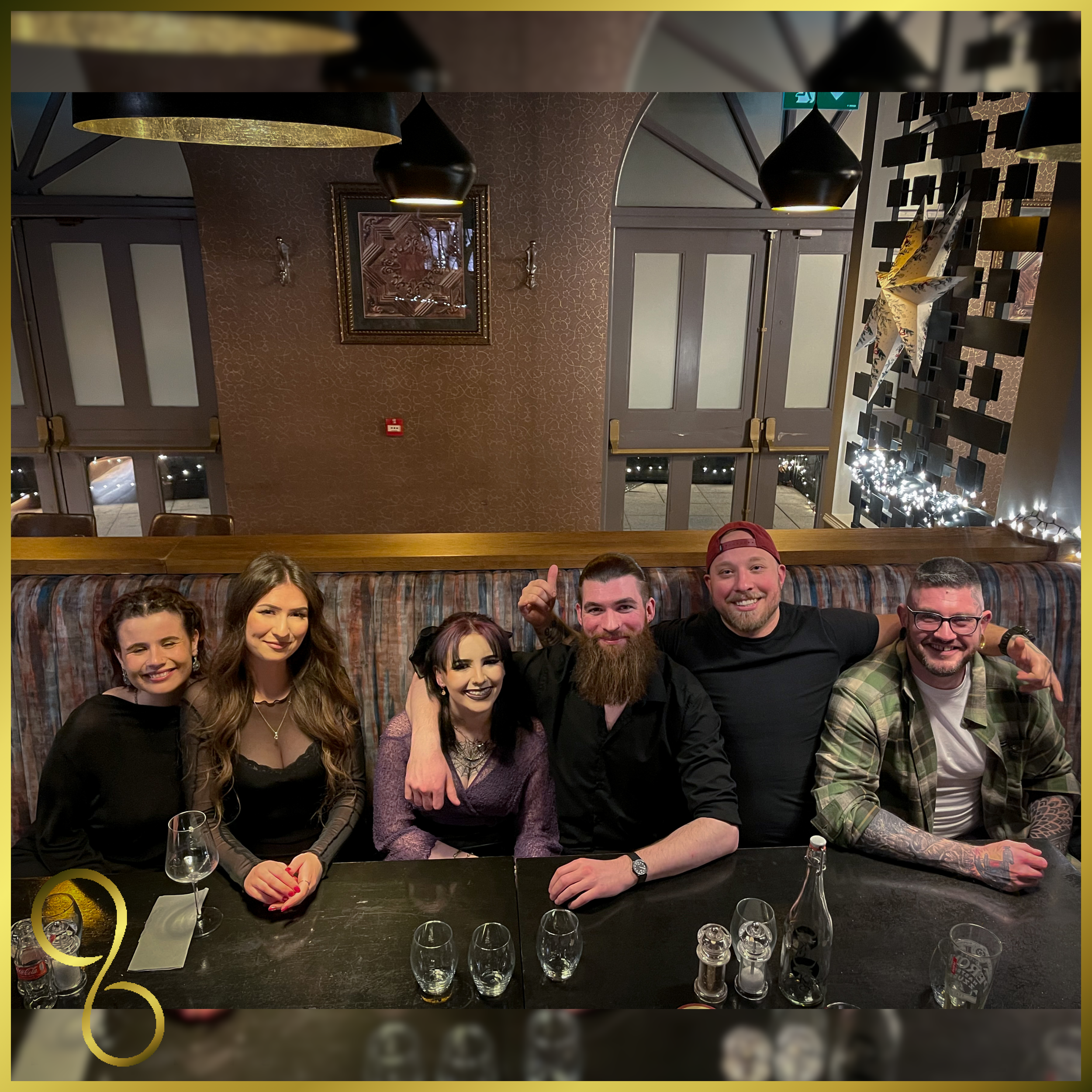 Group of six friends sitting at a restaurant table, smiling and posing for the photo, with drinks and glasses in front of them, decorated for a celebration.
