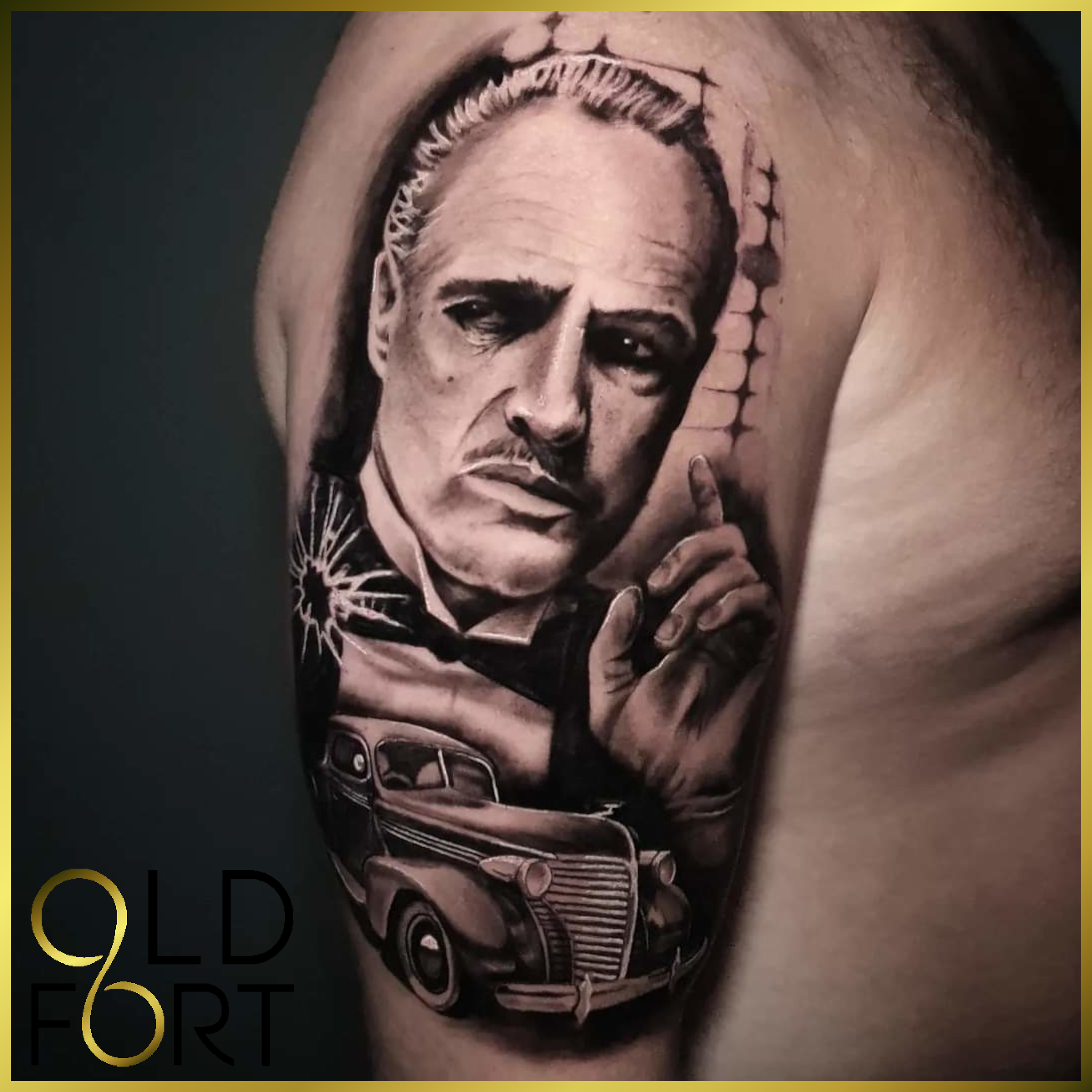 Black and gray tattoo of a man with slicked-back hair, making a gesture with his hand, with an old car below his portrait, on a person's upper arm.