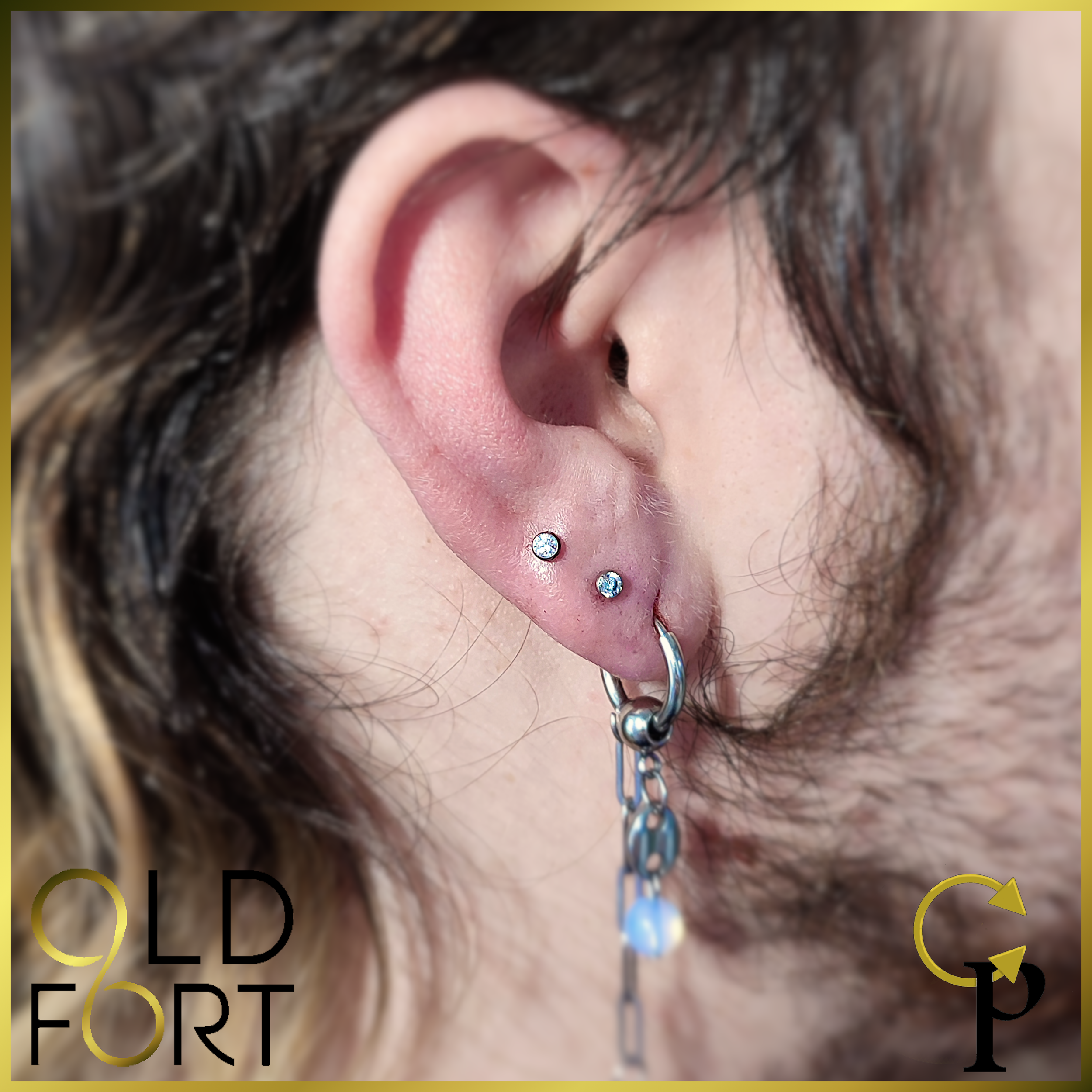 Close-up of a person's ear with multiple piercings, including two small studs and a hoop with a dangling chain and bead.