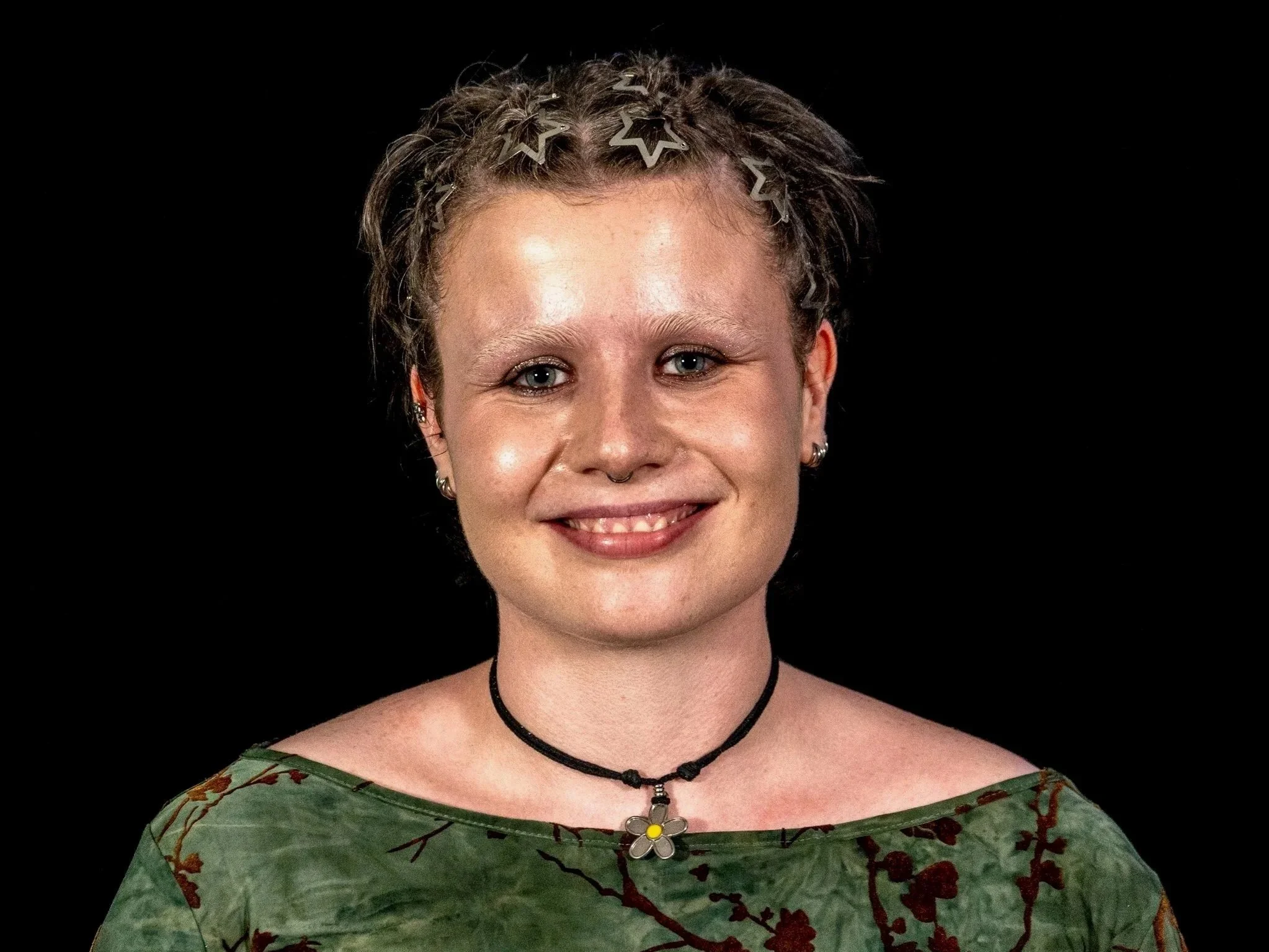 Portrait of a young woman with curly hair adorned with star-shaped hairclips, wearing a floral green top and a black choker with a flower pendant, smiling against a black background.