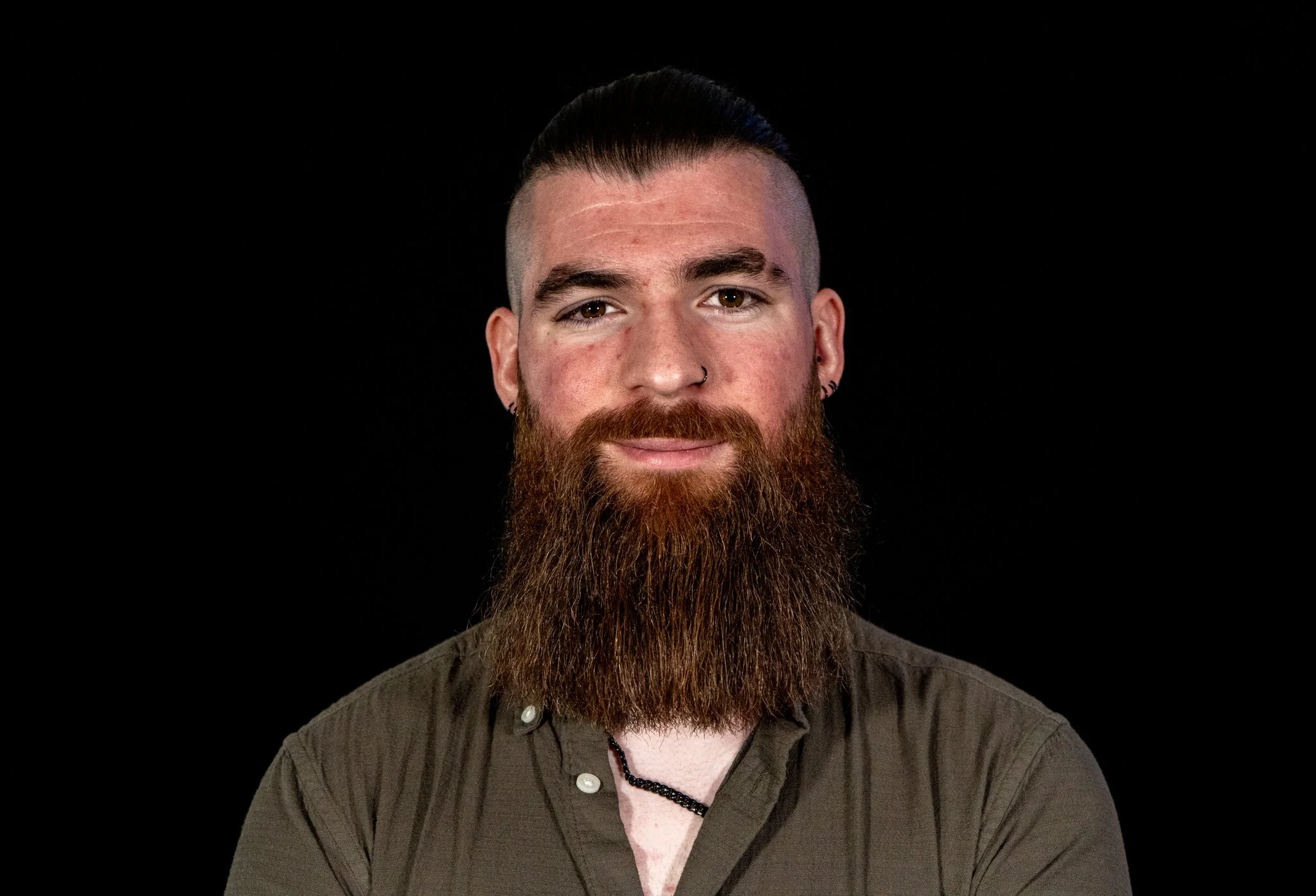 A man with a full beard, dark hair styled with a fade, wearing a nose ring and earrings, dressed in a green button-up shirt against a black background.