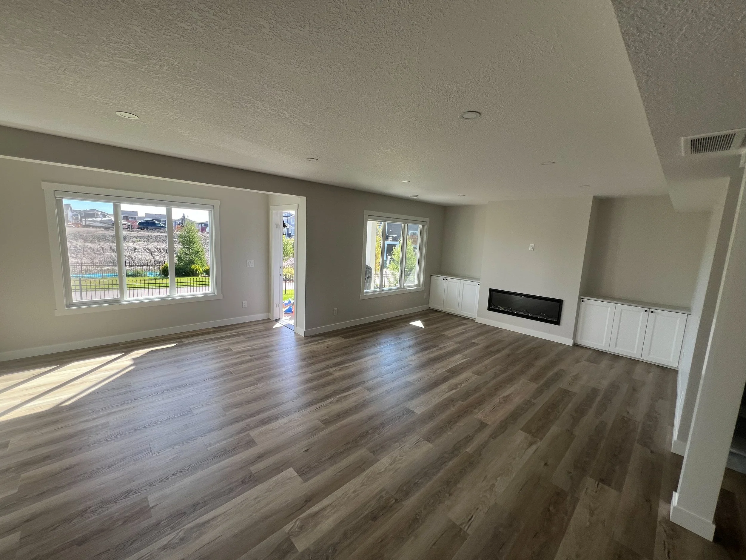 Interior of an empty living room with light wood flooring, large windows, and built-in cabinets with a fireplace.