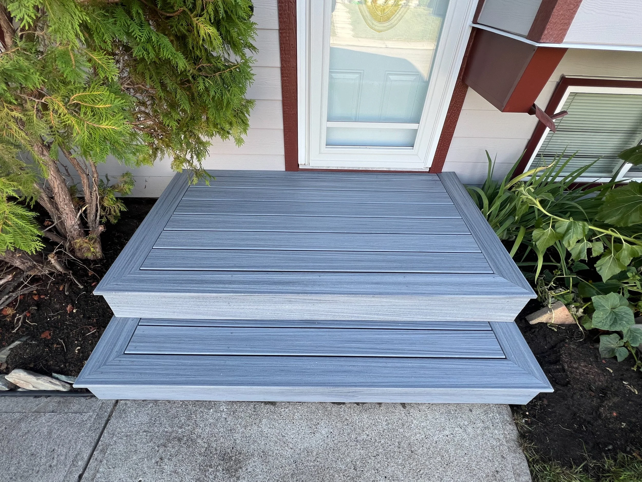 Gray composite decking steps leading to a door, surrounded by greenery and adjacent plants.