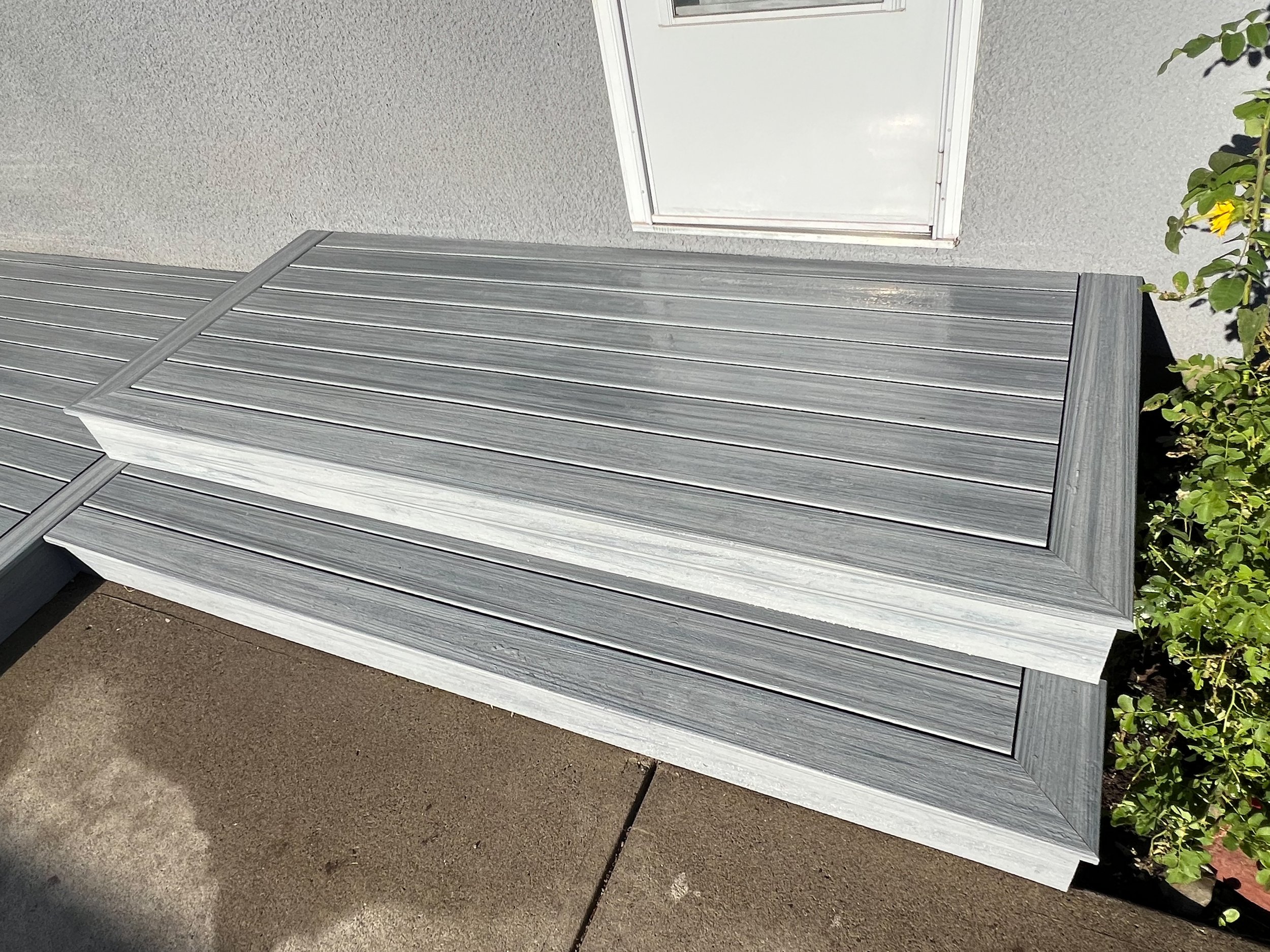 Gray composite deck steps leading to a white door with greenery on the side, in sunlight.