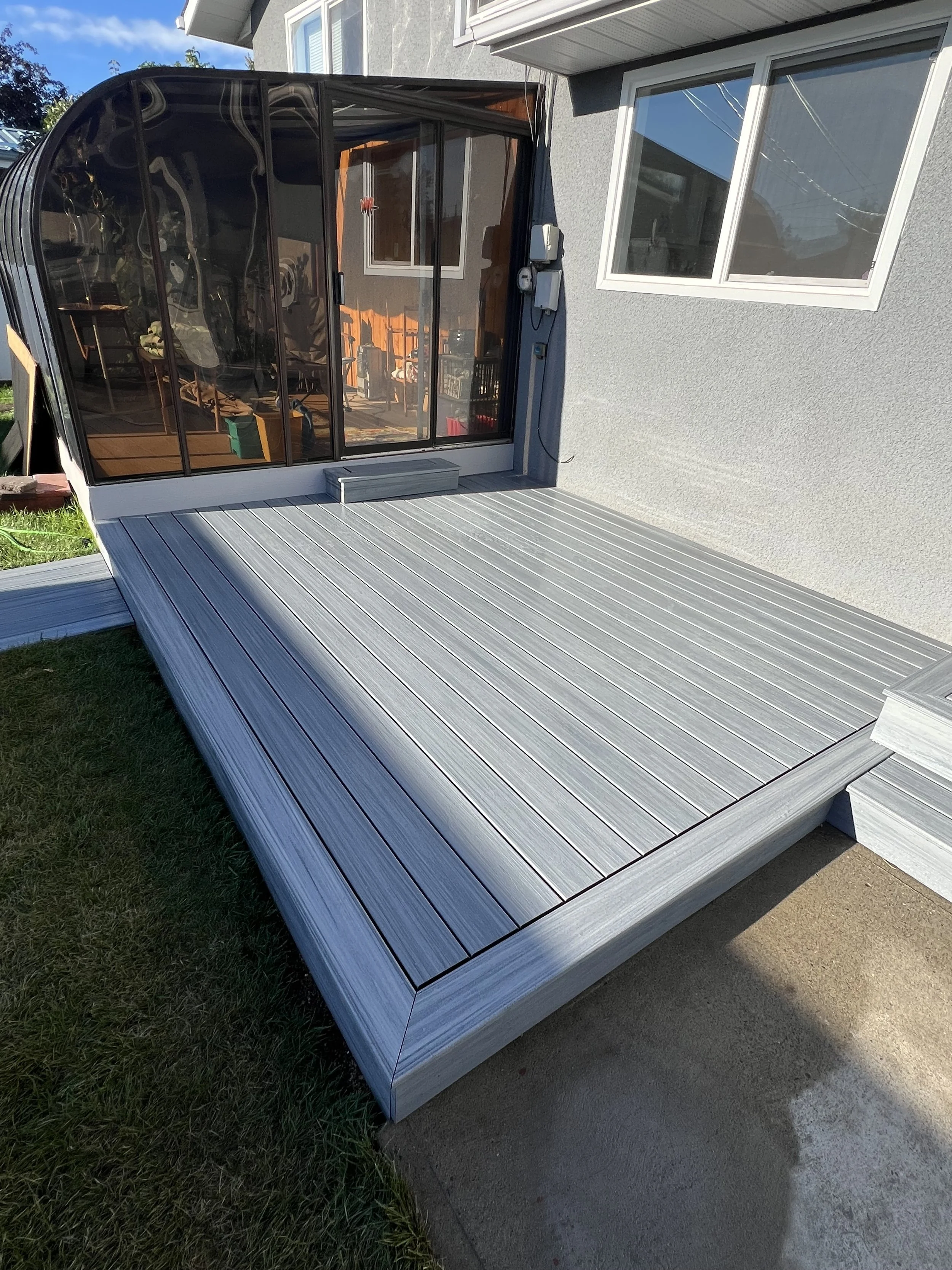 A small deck attached to a house with a sliding glass door and a curved sunroom structure. The deck is made of light gray composite material, adjacent to a grassy lawn and a concrete path.