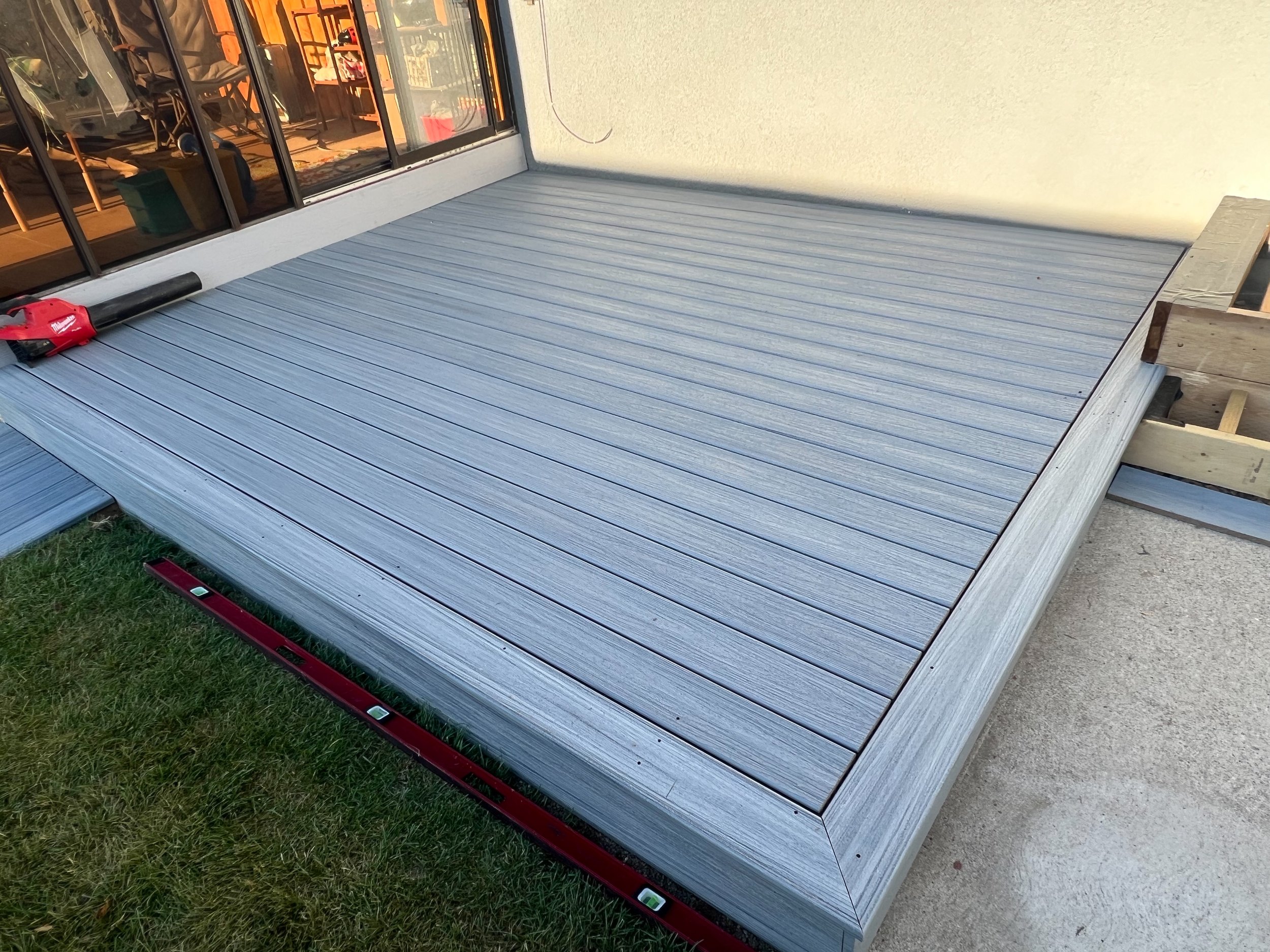 A newly constructed composite deck attached to a building, with a level tool and a leaf blower resting on the surface. The deck is elevated and bordered by a small section of grass and concrete.