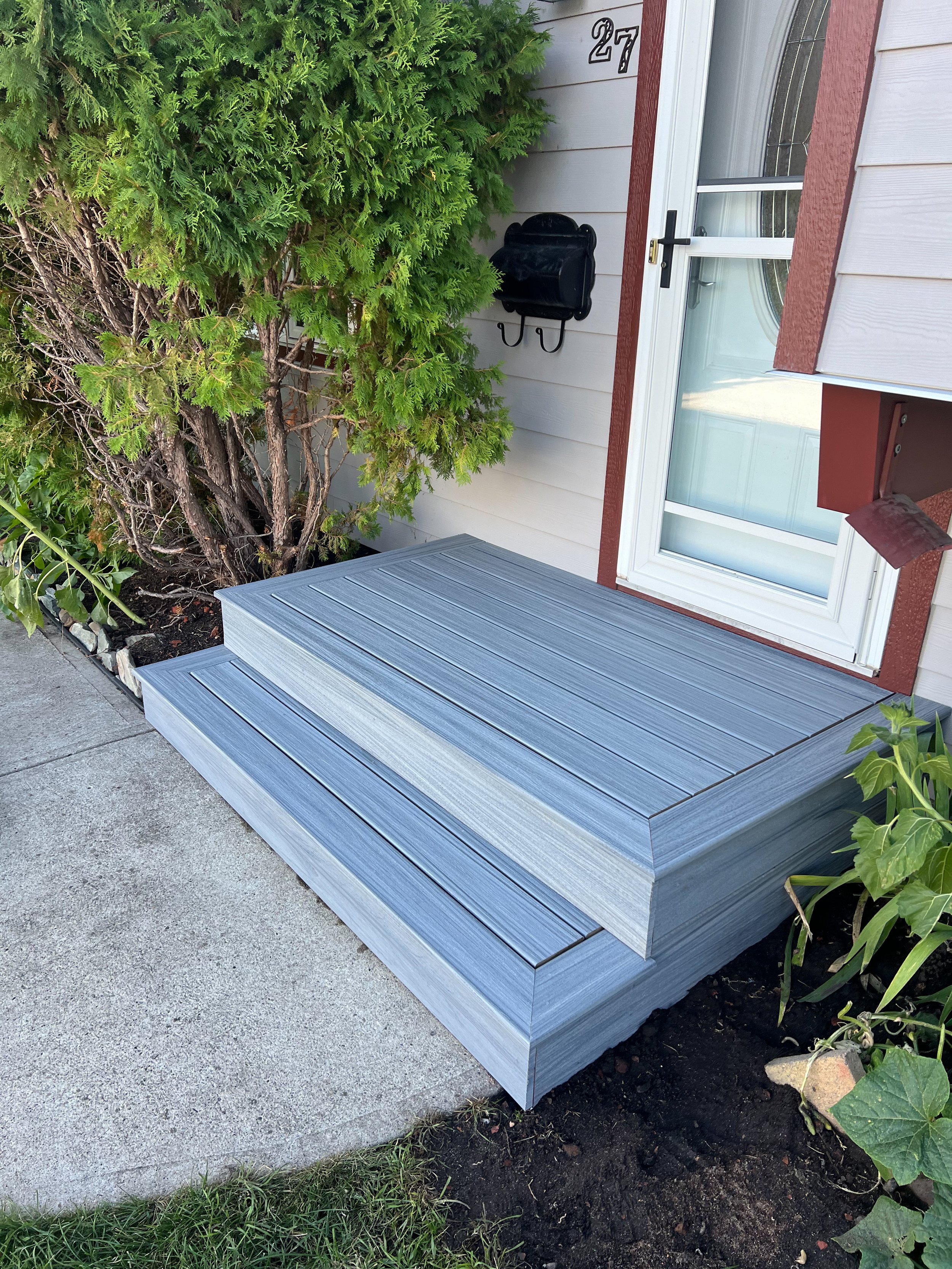 Two-tiered gray steps leading to a front door, surrounded by greenery and landscaping. A white house wall with a door, house number, and mailbox visible.