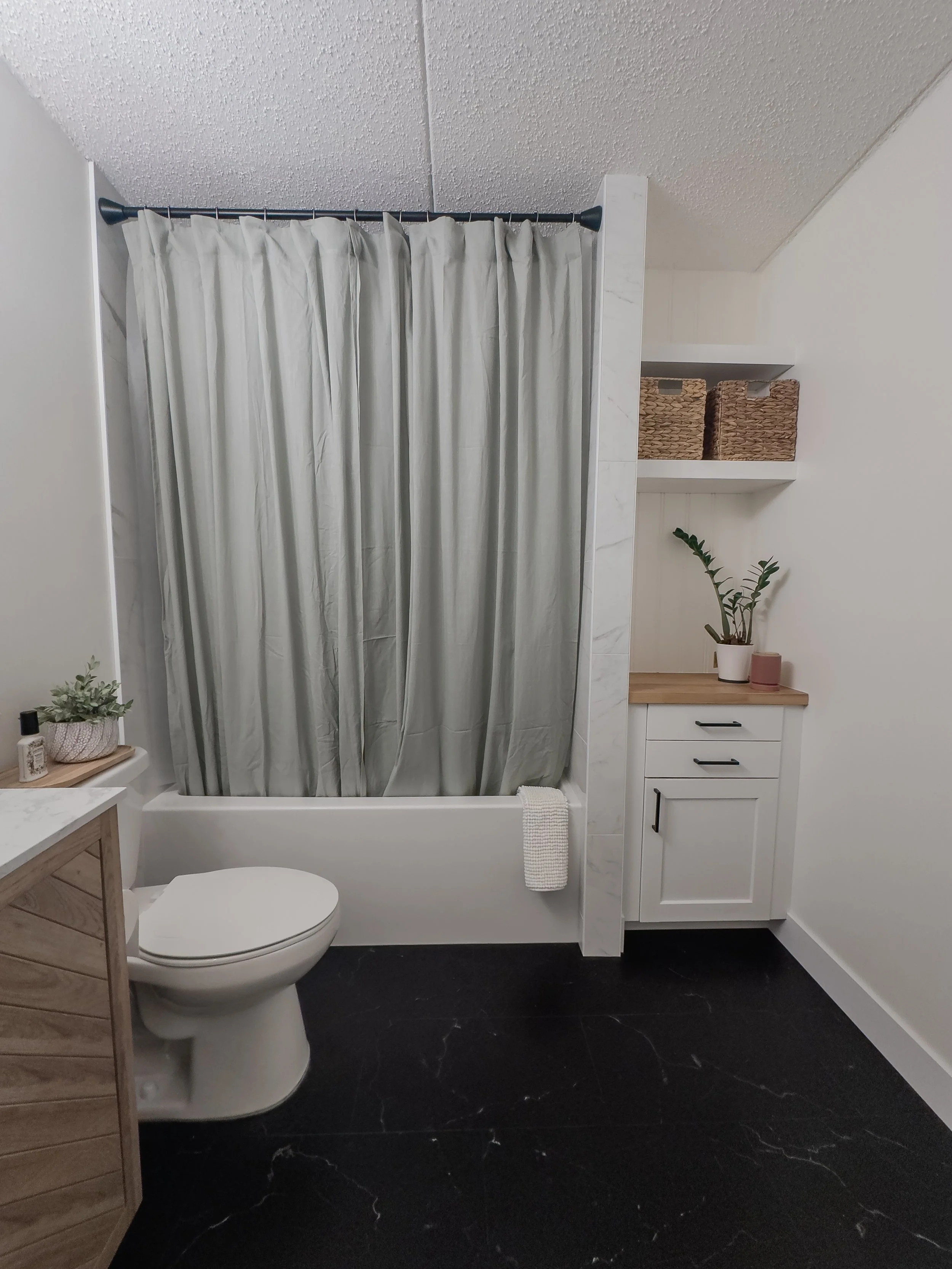 Modern bathroom with a white bathtub and green shower curtain, black floor tiles, white cabinets with potted plants on the counter, and woven baskets on a shelf.