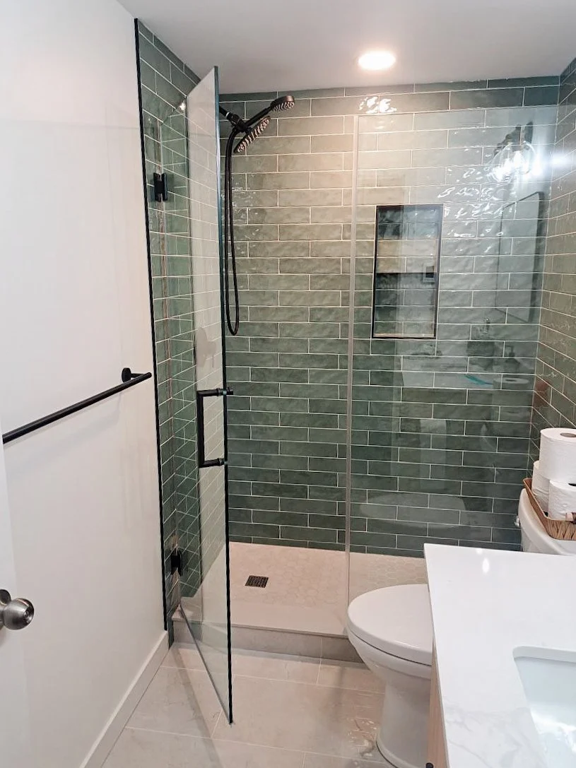 Modern bathroom with green tiled shower, glass door, and white fixtures; a basket with toilet paper is on the counter.