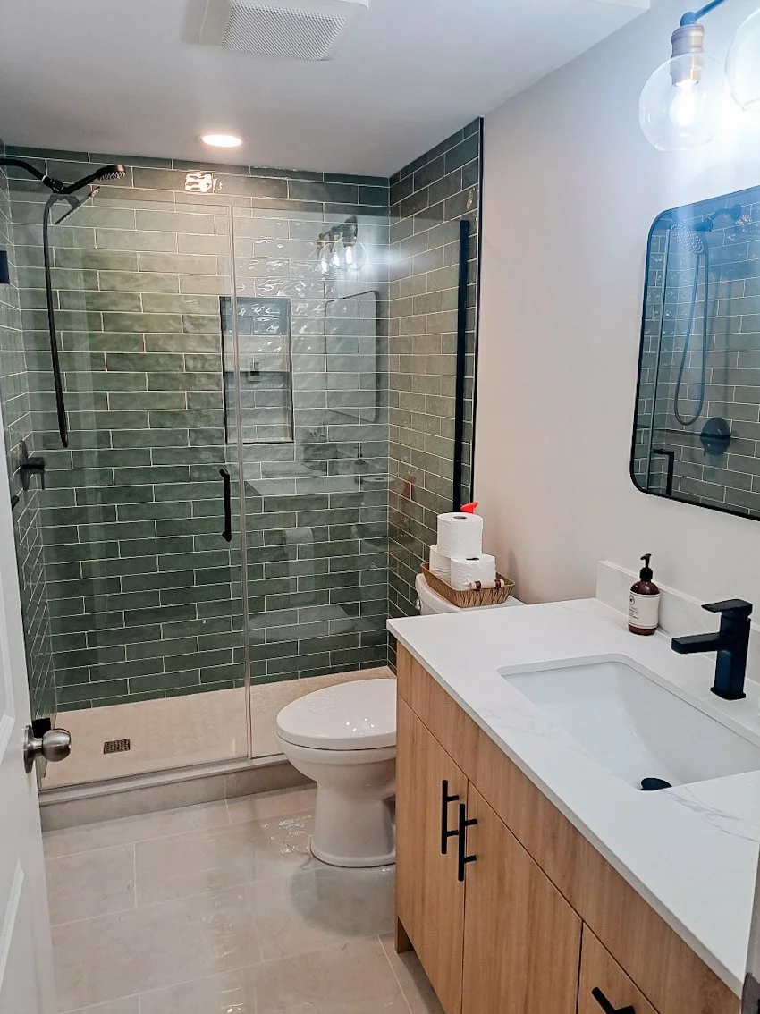 Modern bathroom with green-tiled shower, glass door, wooden vanity, white sink, and black fixtures.
