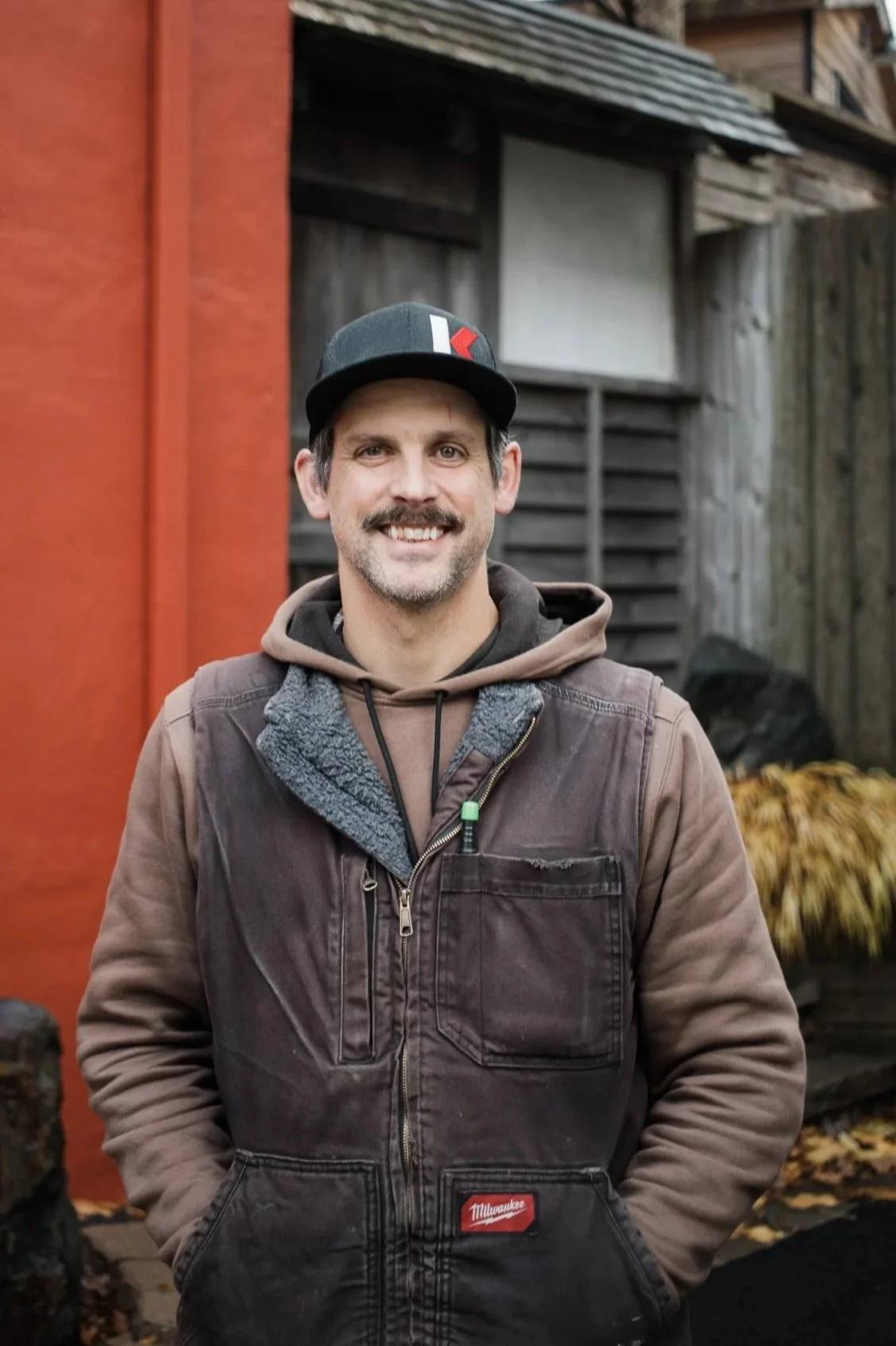 A smiling man wearing a black baseball cap, brown hoodie, and brown jacket standing outdoors with a rustic wooden and metal building in the background.