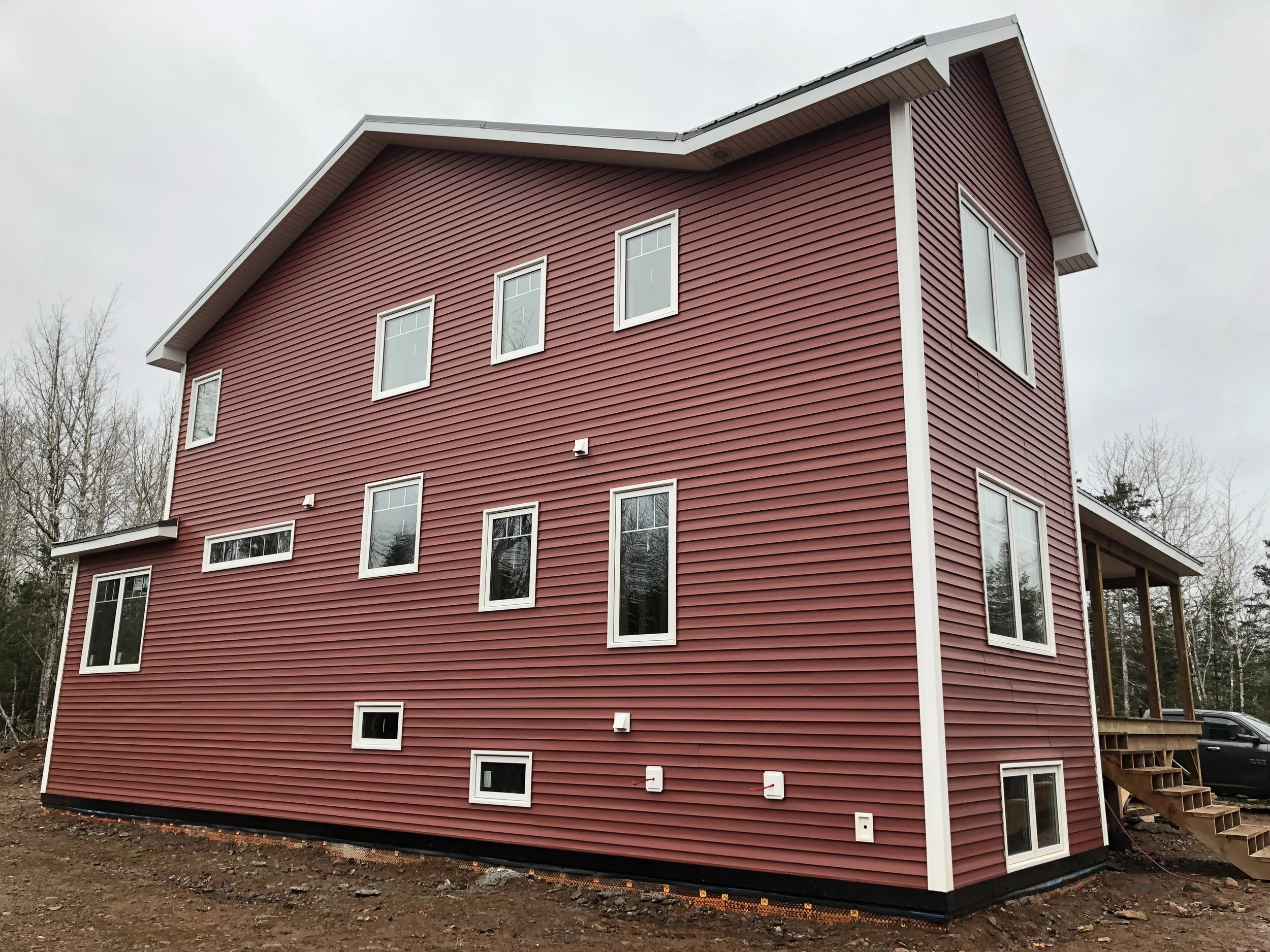 A multi-story house with red siding and multiple white-framed windows under construction. The house is on a foundation with dirt and some construction materials around. The sky is overcast.
