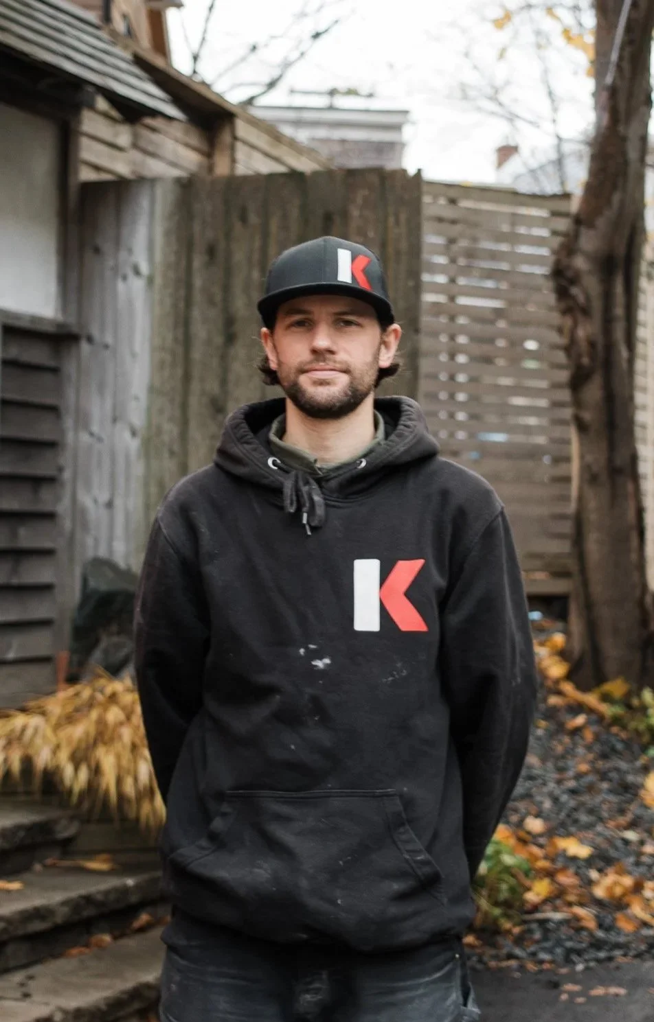 A man with dark hair and beard wearing a black hoodie and cap with a red and white logo stands outdoors in front of a wooden fence and trees with autumn leaves.