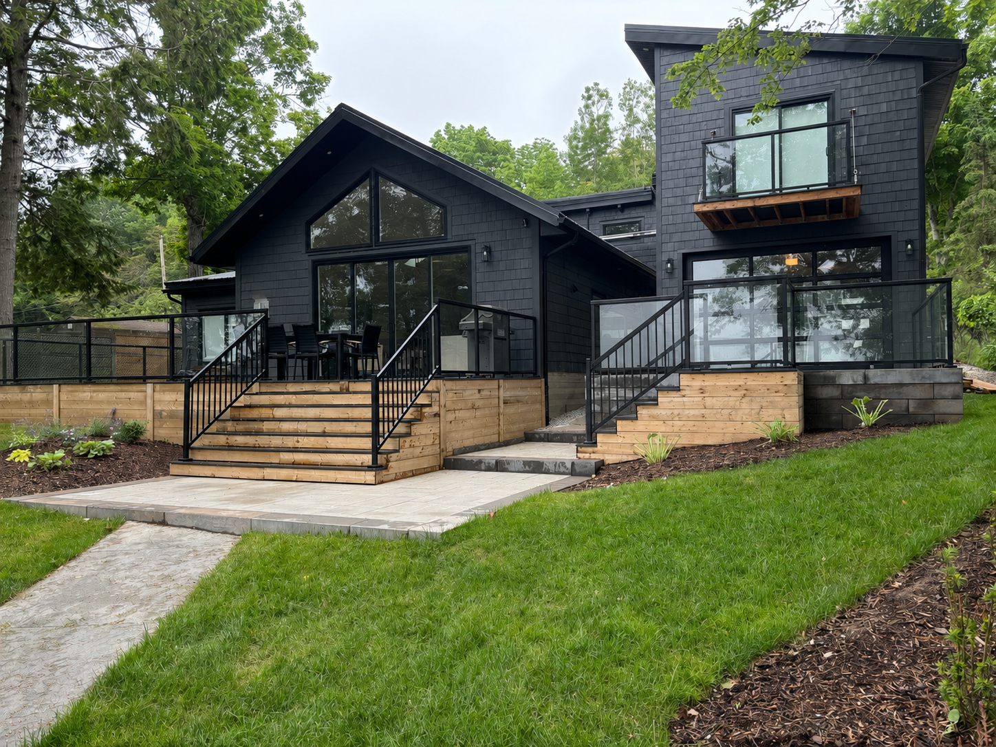 Modern black house with multiple decks, large glass windows, and a metal railing. The house is surrounded by green grass, trees, and landscaped gardens.