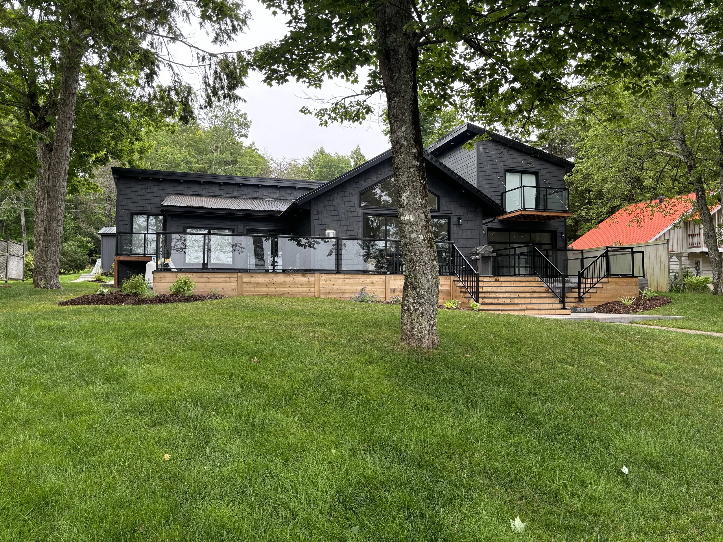 A modern black house with large windows, a deck, and stairs in a backyard with green grass, surrounded by tall trees and a partly cloudy sky.