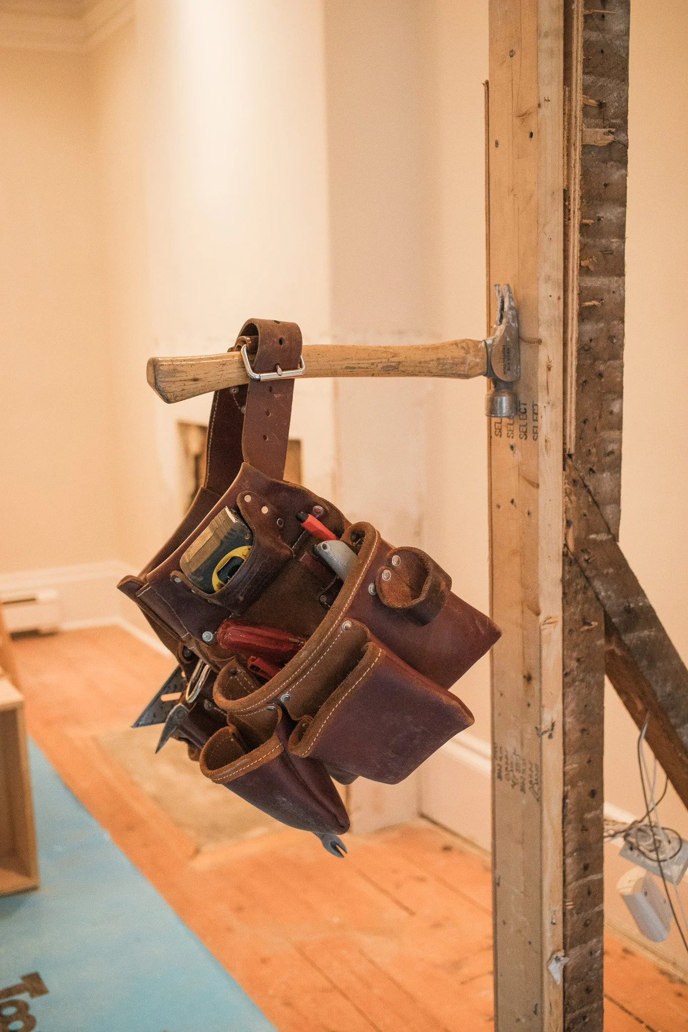Construction tools including a hammer and a tool belt hanging from a wooden frame during interior renovation.