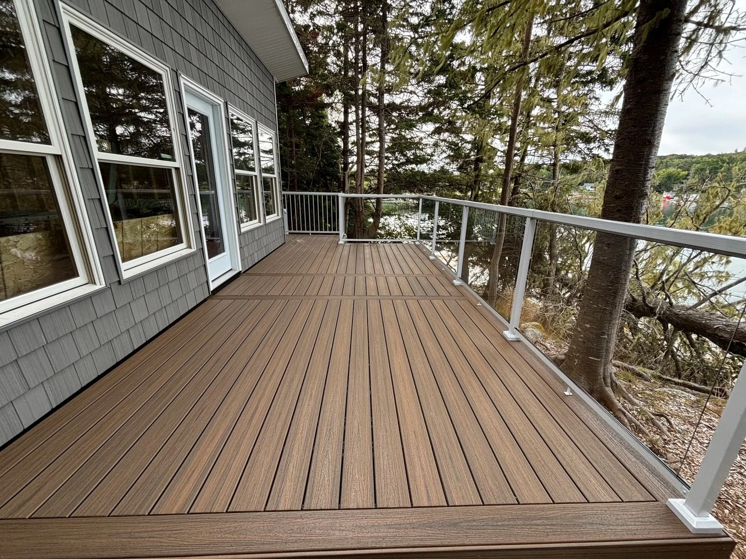 A wooden balcony with a glass railing surrounded by trees and a view of a river or water body in the distance.