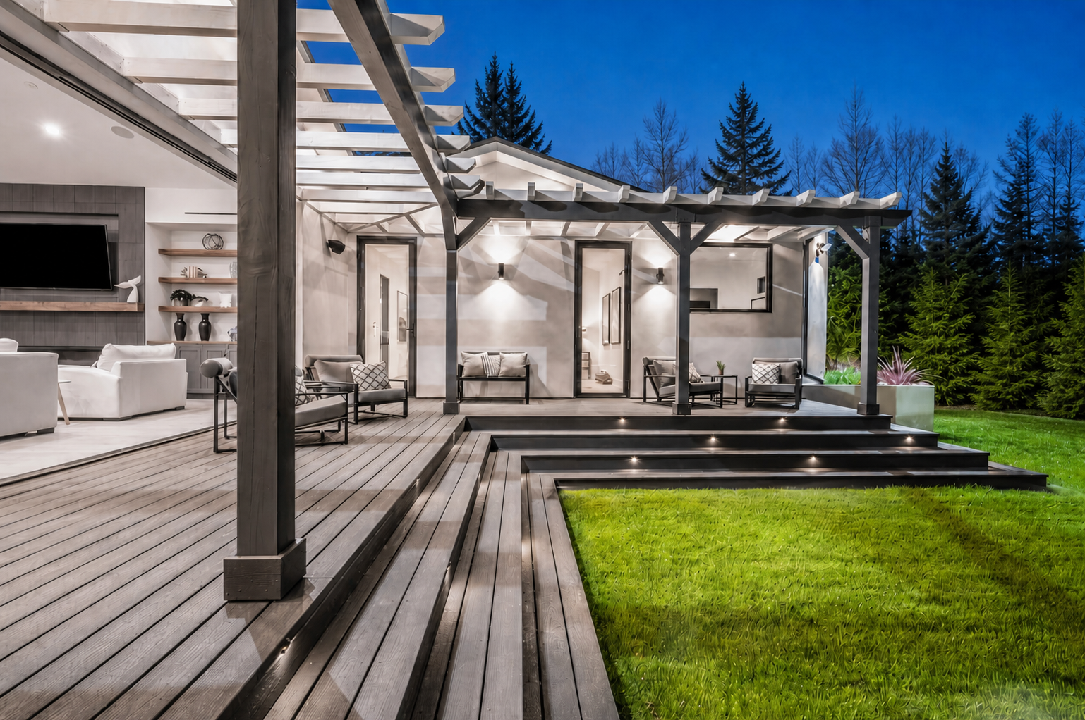 Night view of a modern house's outdoor patio with wooden decking, seating area, and steps leading down to a grassy lawn, surrounded by tall pine trees.