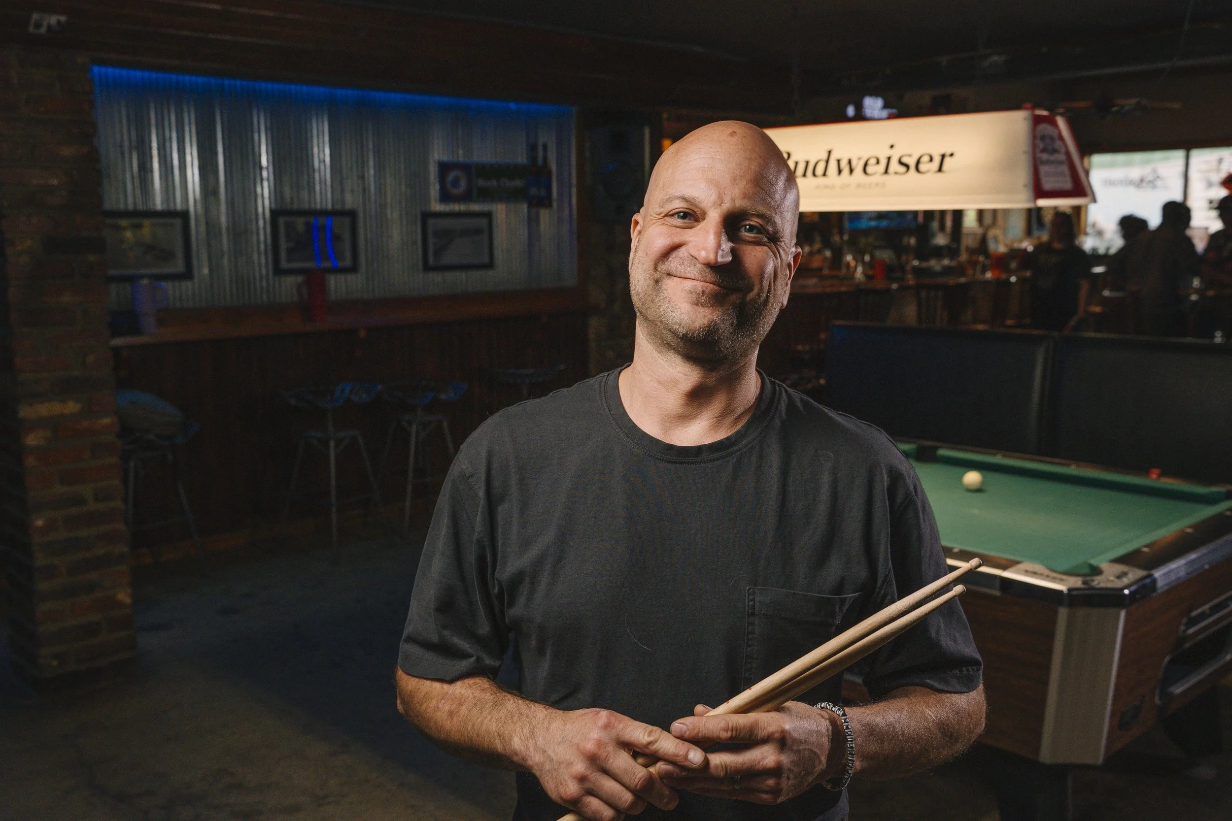 A smiling man holding pool cues in a dimly lit bar with a pool table and a Budweiser sign in the background.